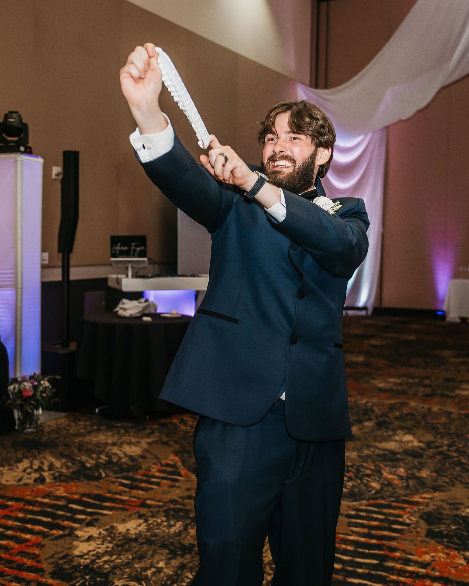 Groom in navy suit holds up garter at wedding reception, smiling triumphantly on patterned ballroom floor.