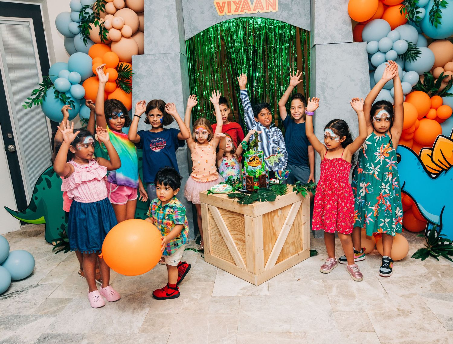 Children celebrating a dinosaur-themed birthday party with colorful balloons and green tinsel backdrop.