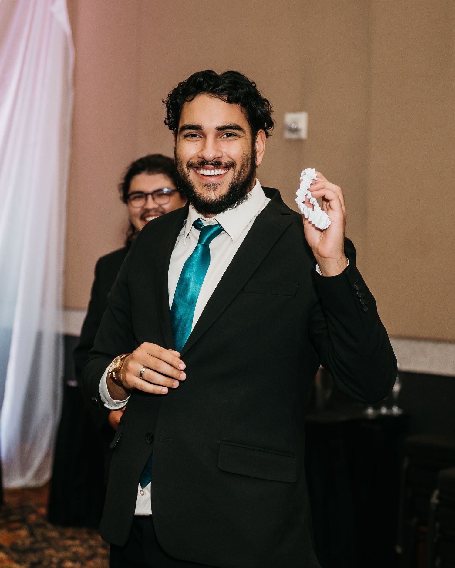 Smiling man in black suit with teal tie holding small item at formal event.