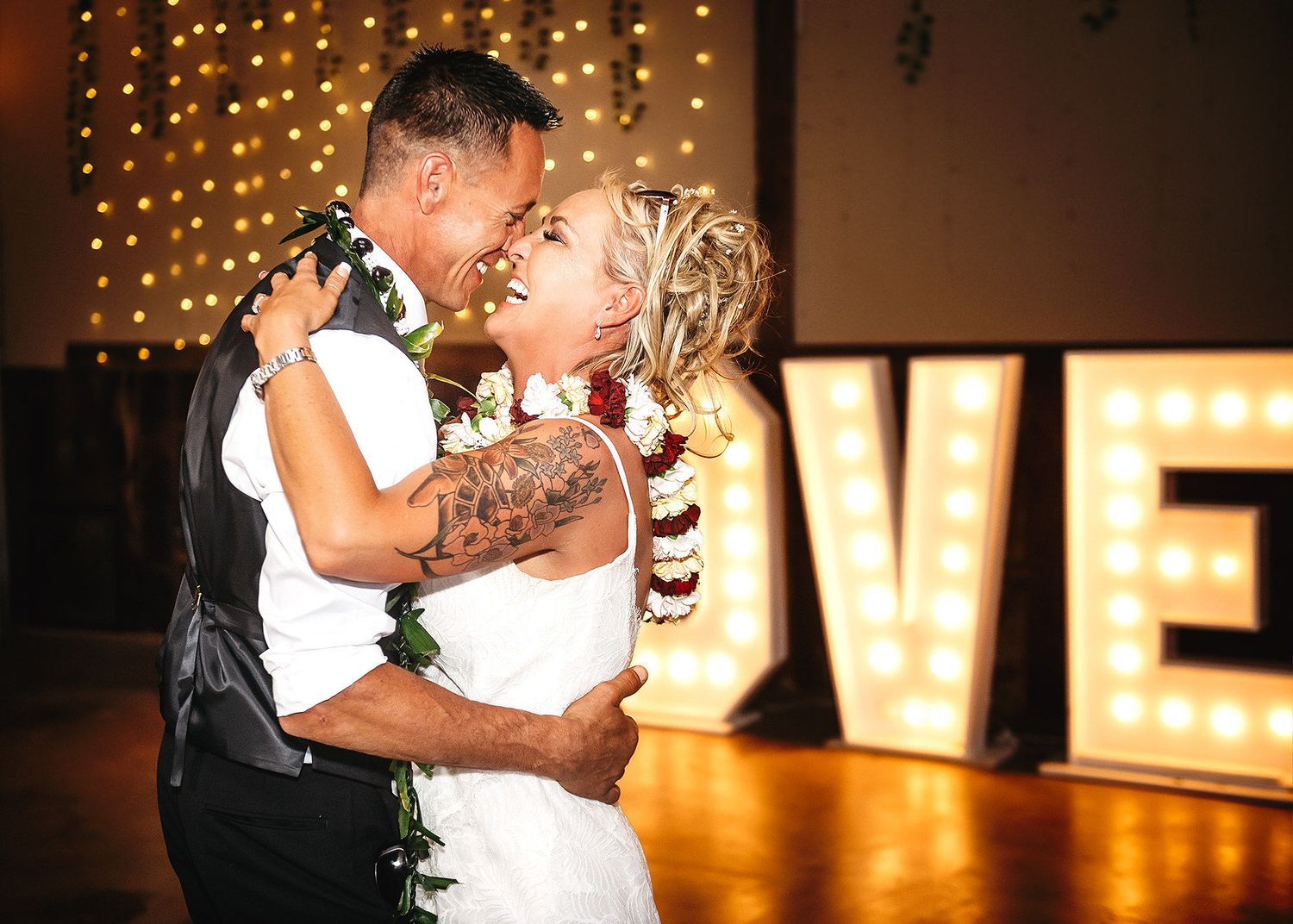 Bride and groom share first dance kiss in front of illuminated LOVE letters at romantic wedding reception.