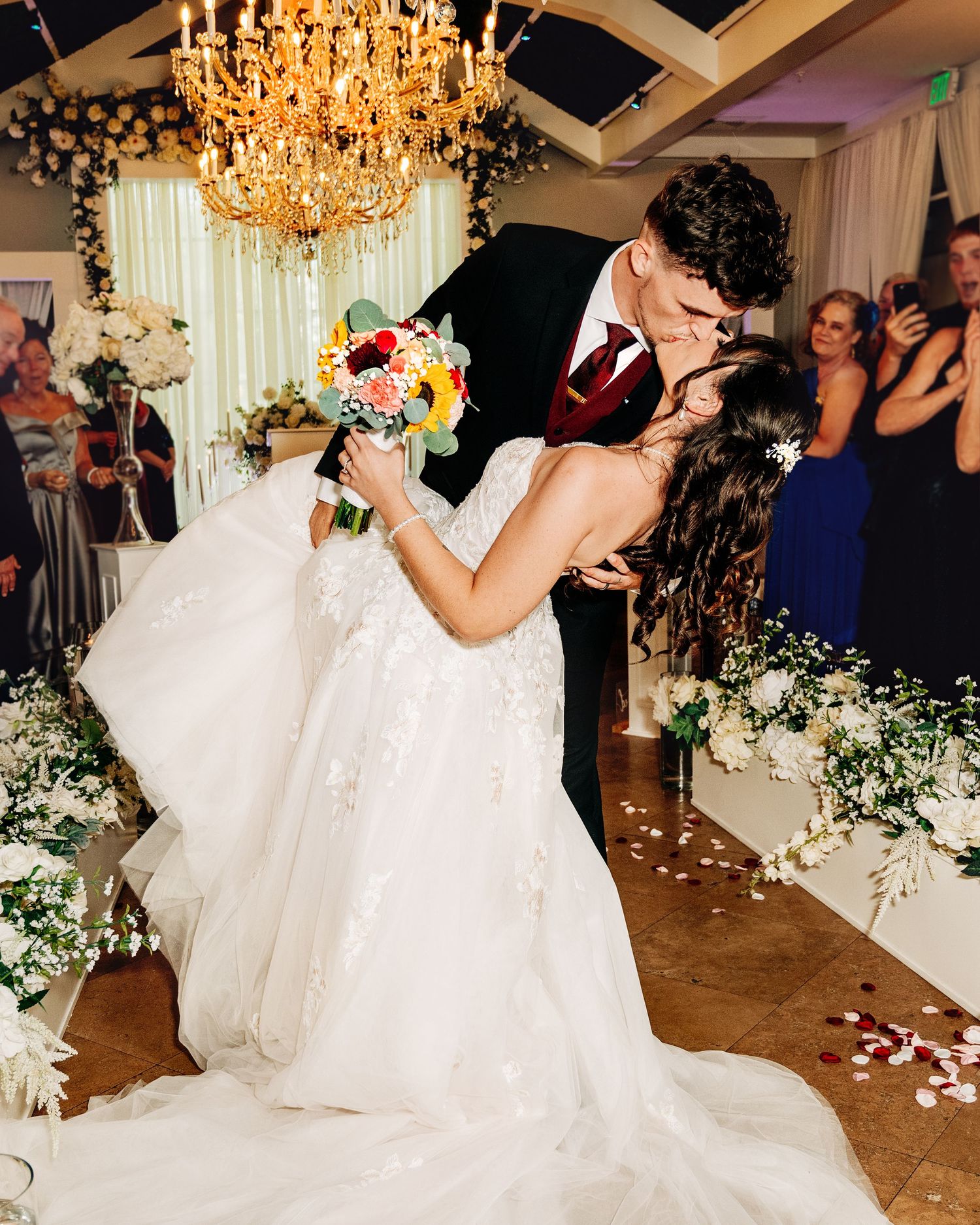 Bride and groom share a romantic dip kiss at their wedding ceremony, surrounded by white floral arrangements and guests.