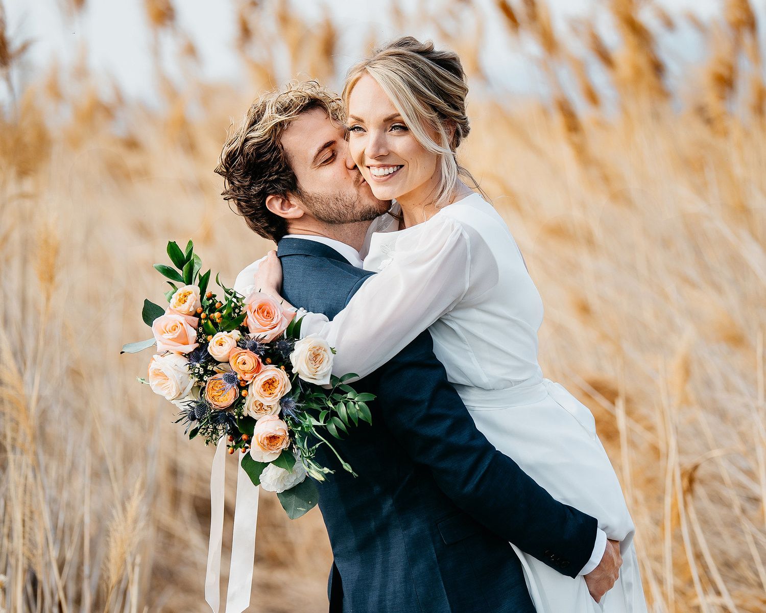 Groom kissing bride on cheek while holding her in a golden wheat field, bride holding peach rose bouquet.