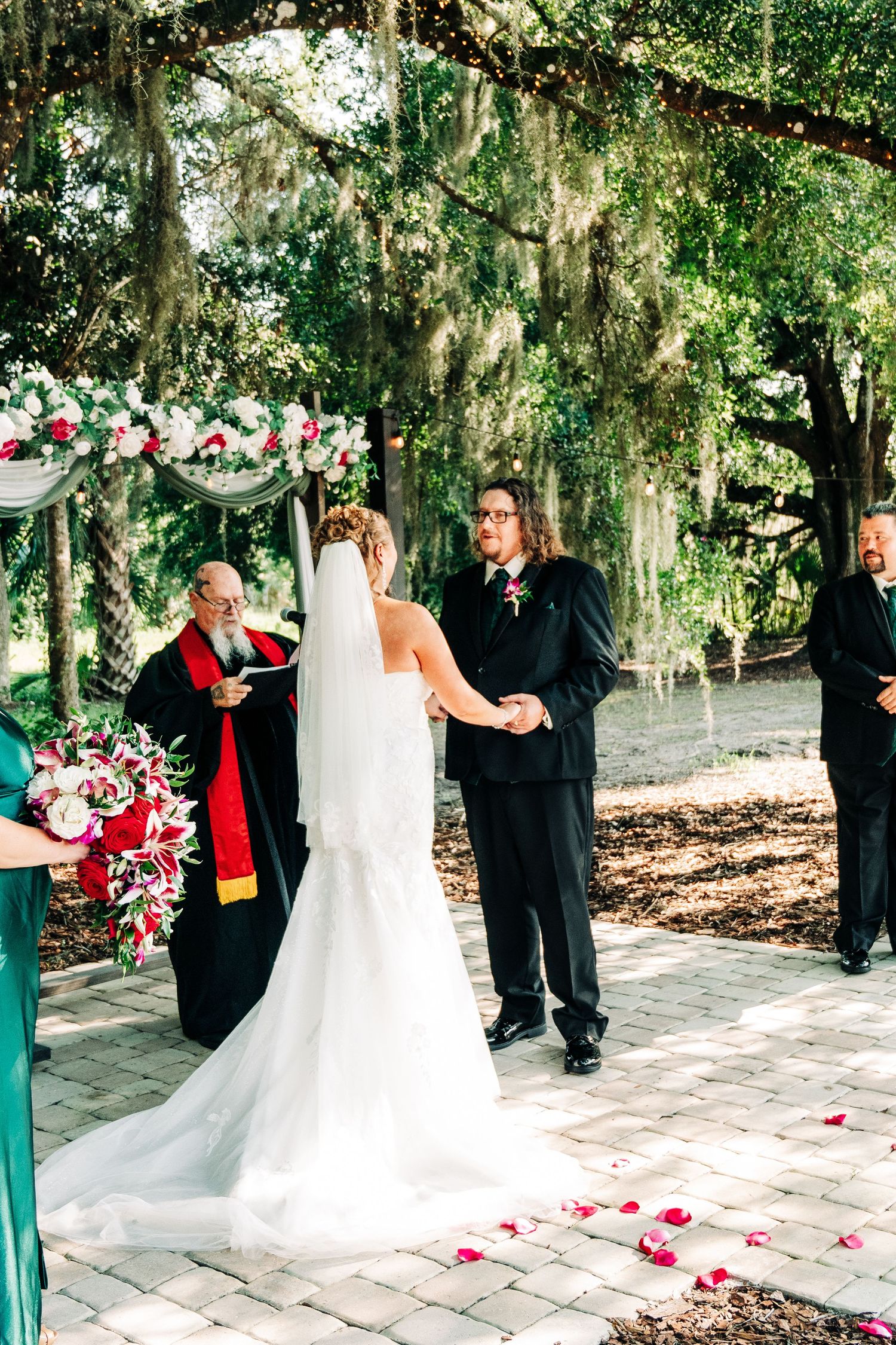 Outdoor wedding ceremony under moss-draped oak trees, bride and groom holding hands before officiant with red stole.
