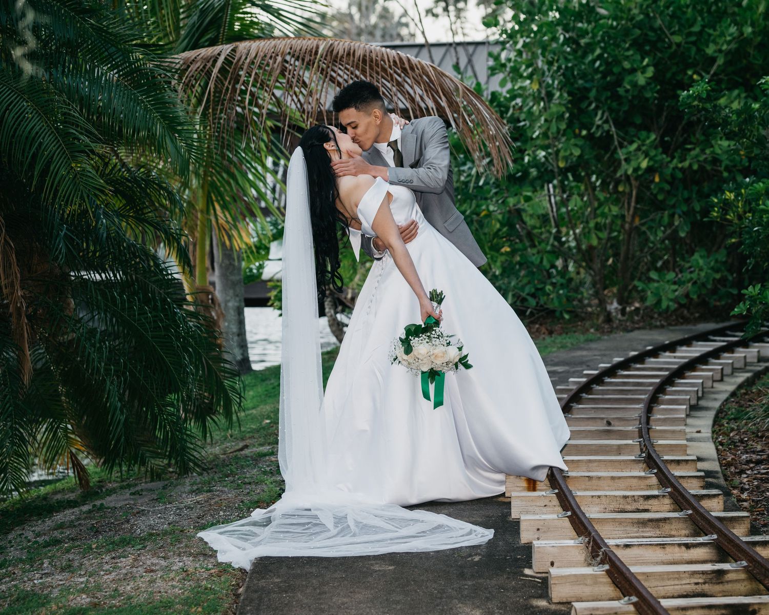 Bride and groom share a romantic dip kiss near railroad tracks surrounded by lush tropical greenery.