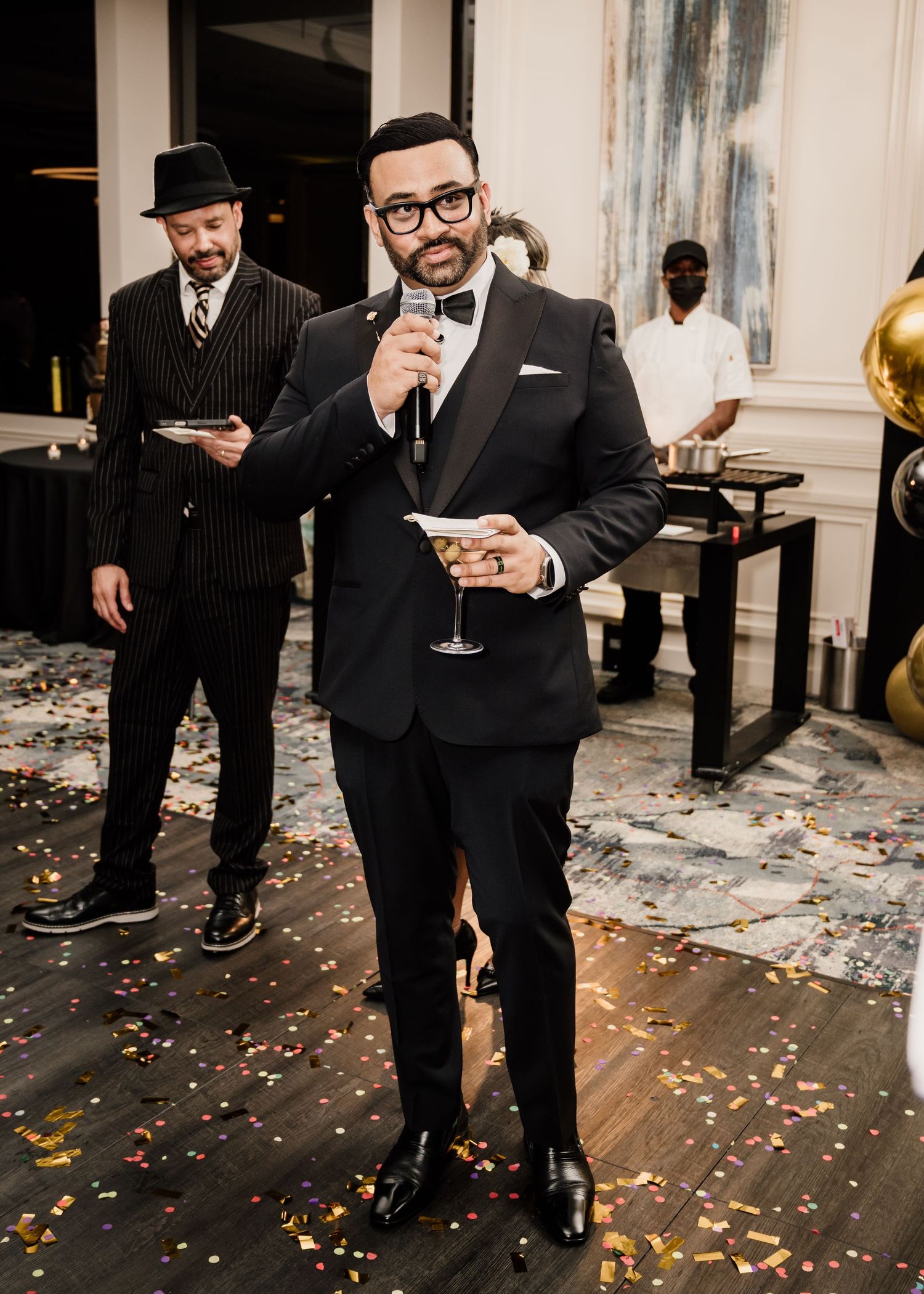 Elegant man in black tuxedo holding champagne glass at formal party with confetti on floor.
