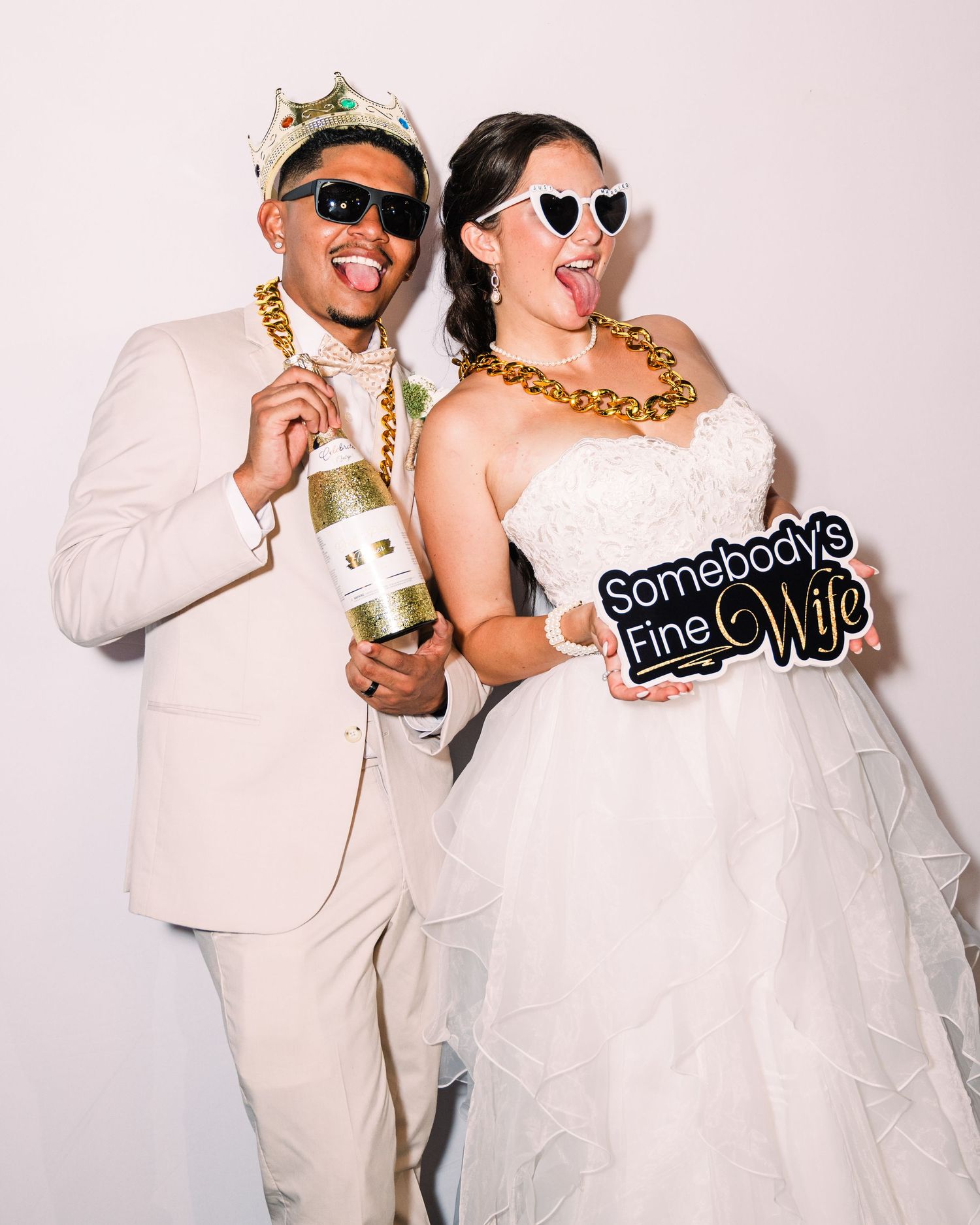 Newlyweds pose in a fun photo booth, wearing oversized sunglasses and gold chains, holding champagne and a humorous wife sign.