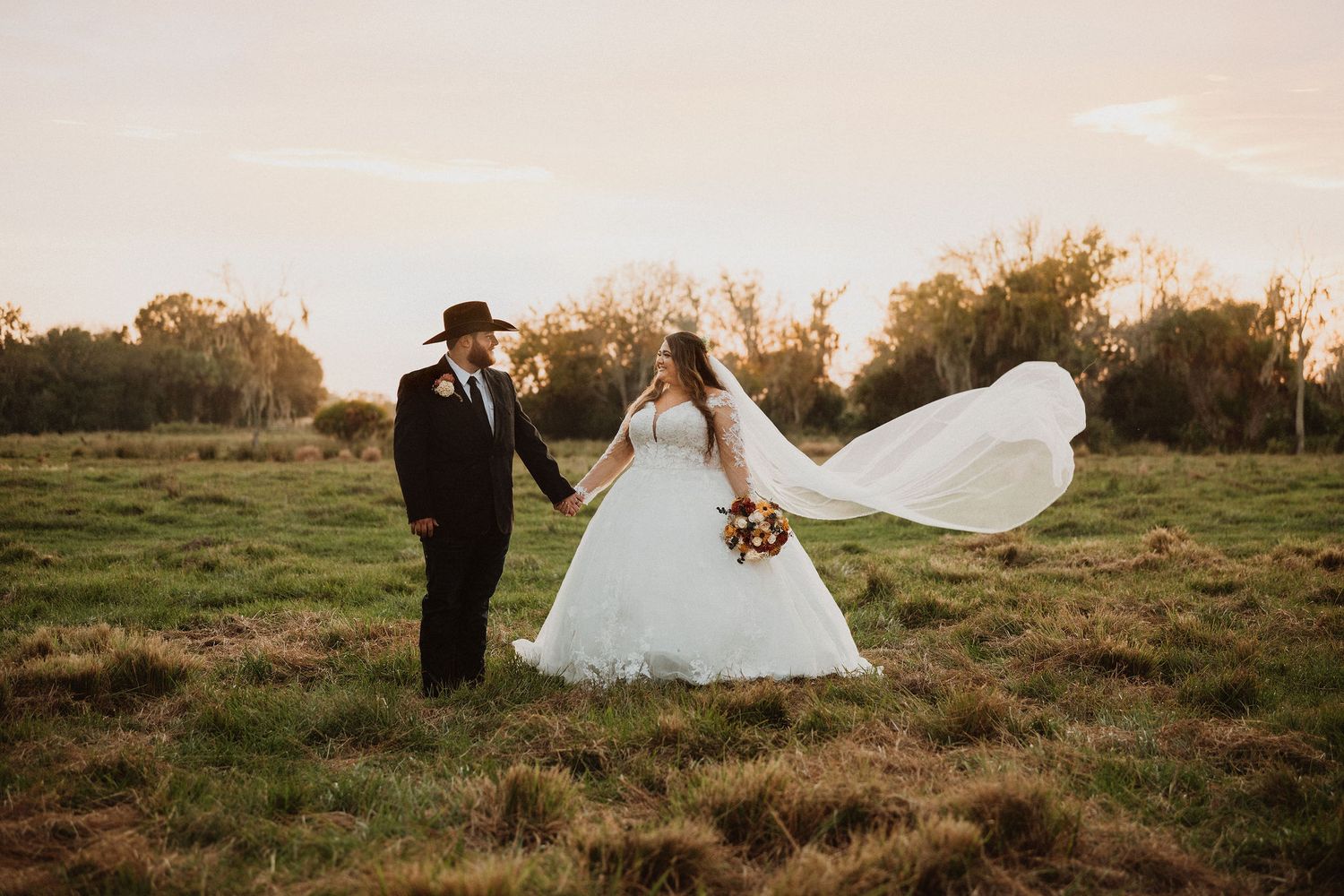 Bride and groom holding hands in a sunlit field, her veil flowing in the wind, groom in black suit and cowboy hat.