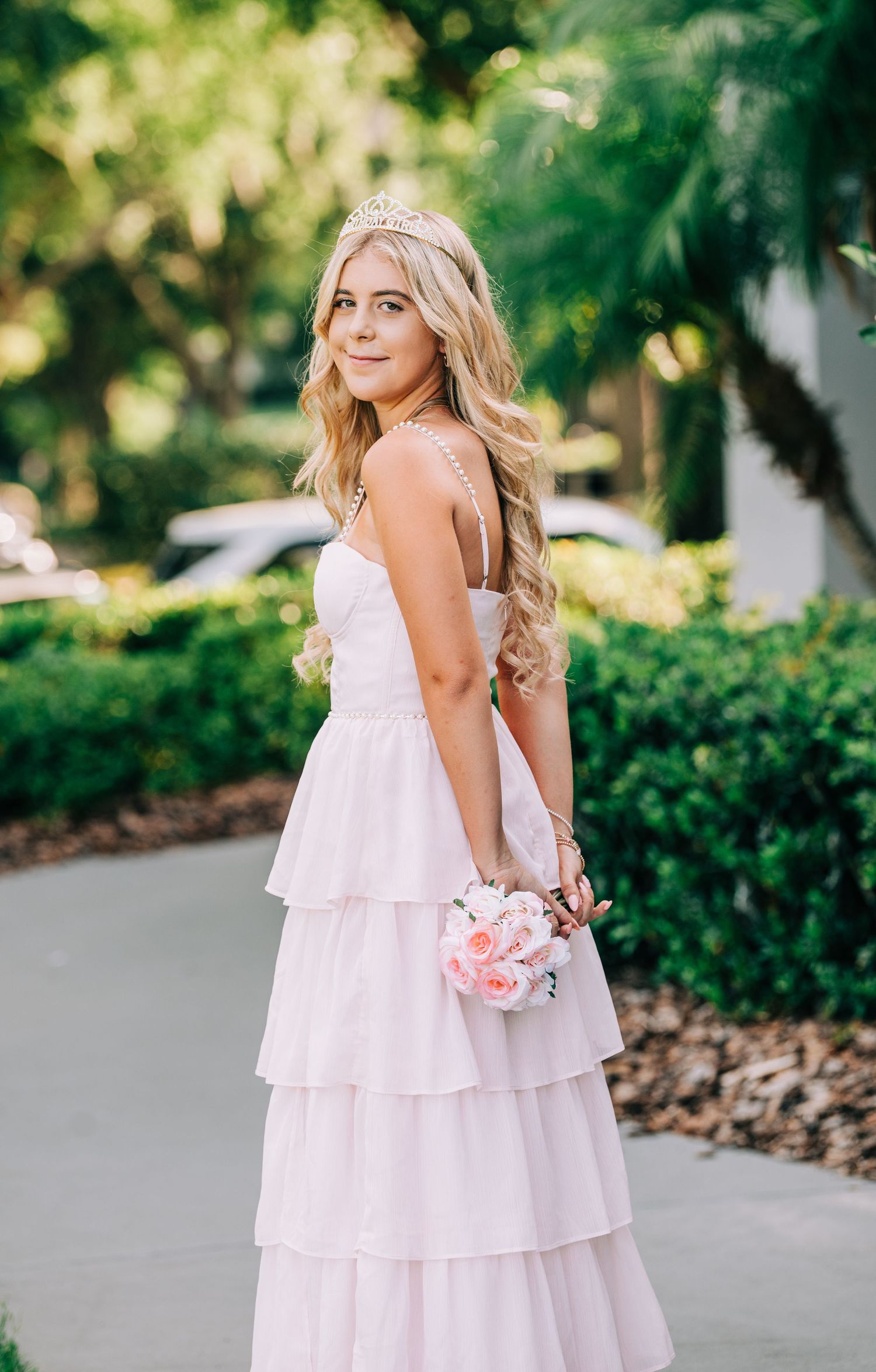 Young blonde woman in a pink tiered formal gown wearing a crown and holding a bouquet outdoors.