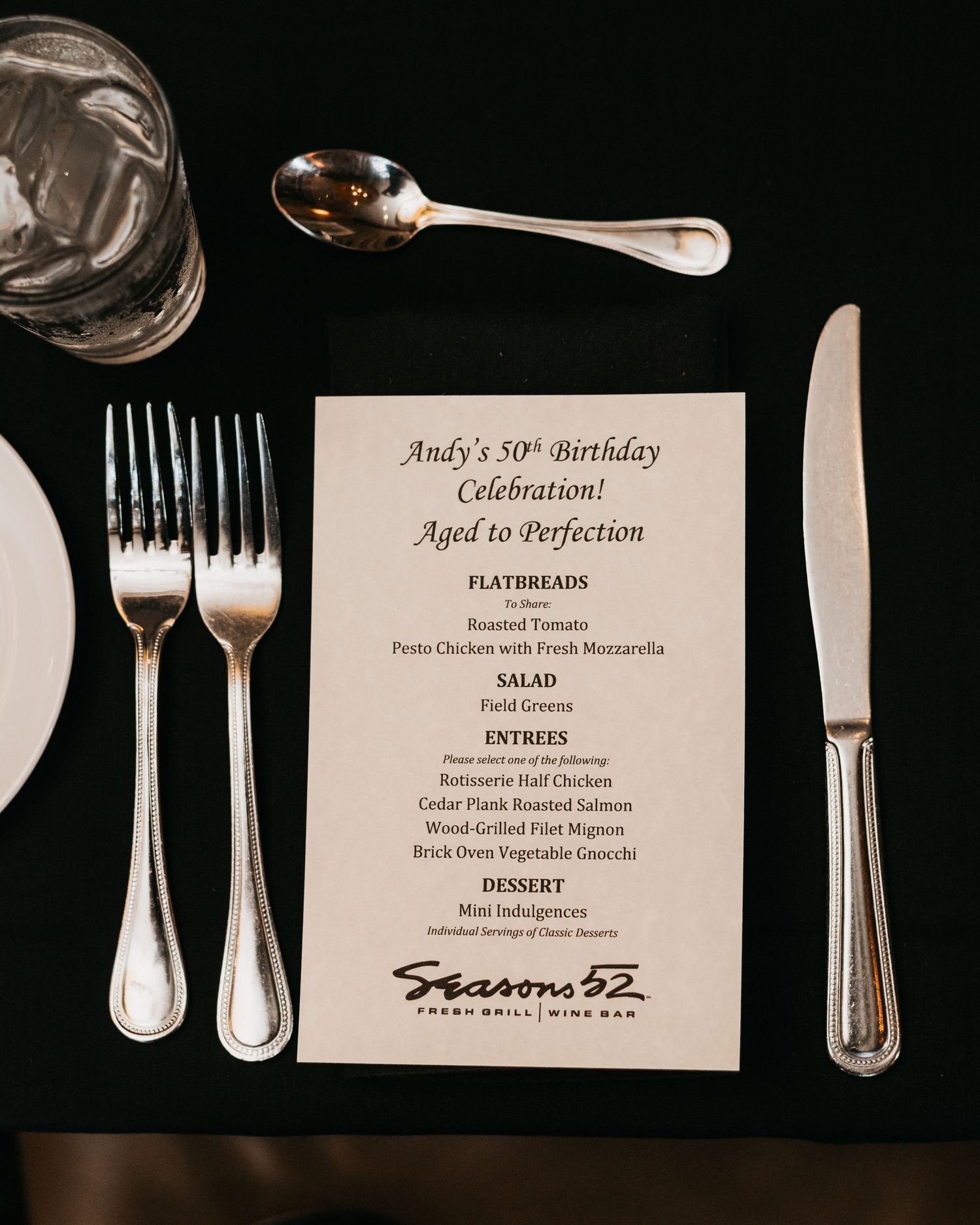 Elegant place setting at Seasons 52 featuring a birthday menu for Andy's 50th celebration with silverware on black tablecloth.