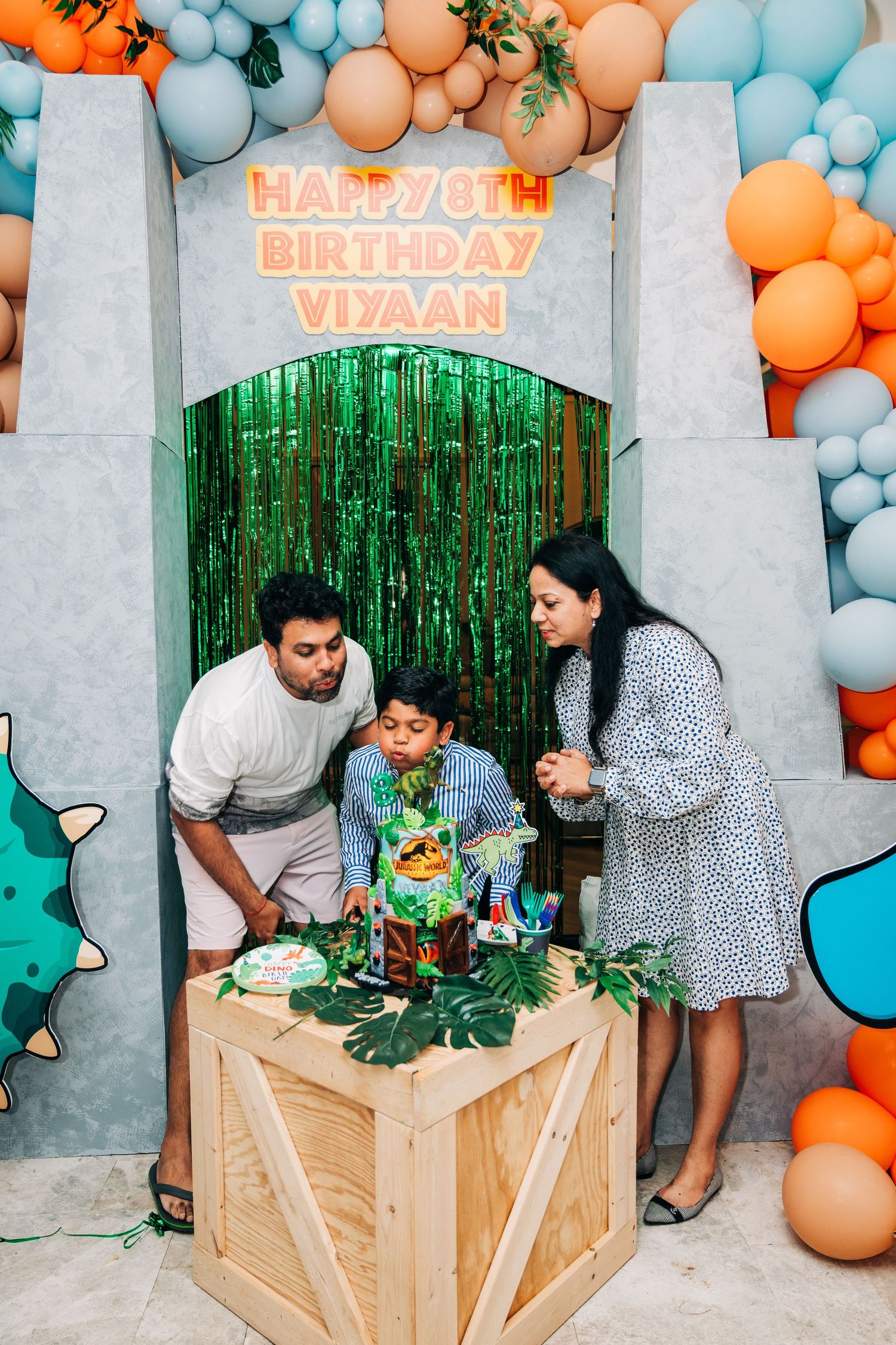 Family celebrating Viyaan's 8th birthday with dinosaur cake, orange and teal balloon arch decor.