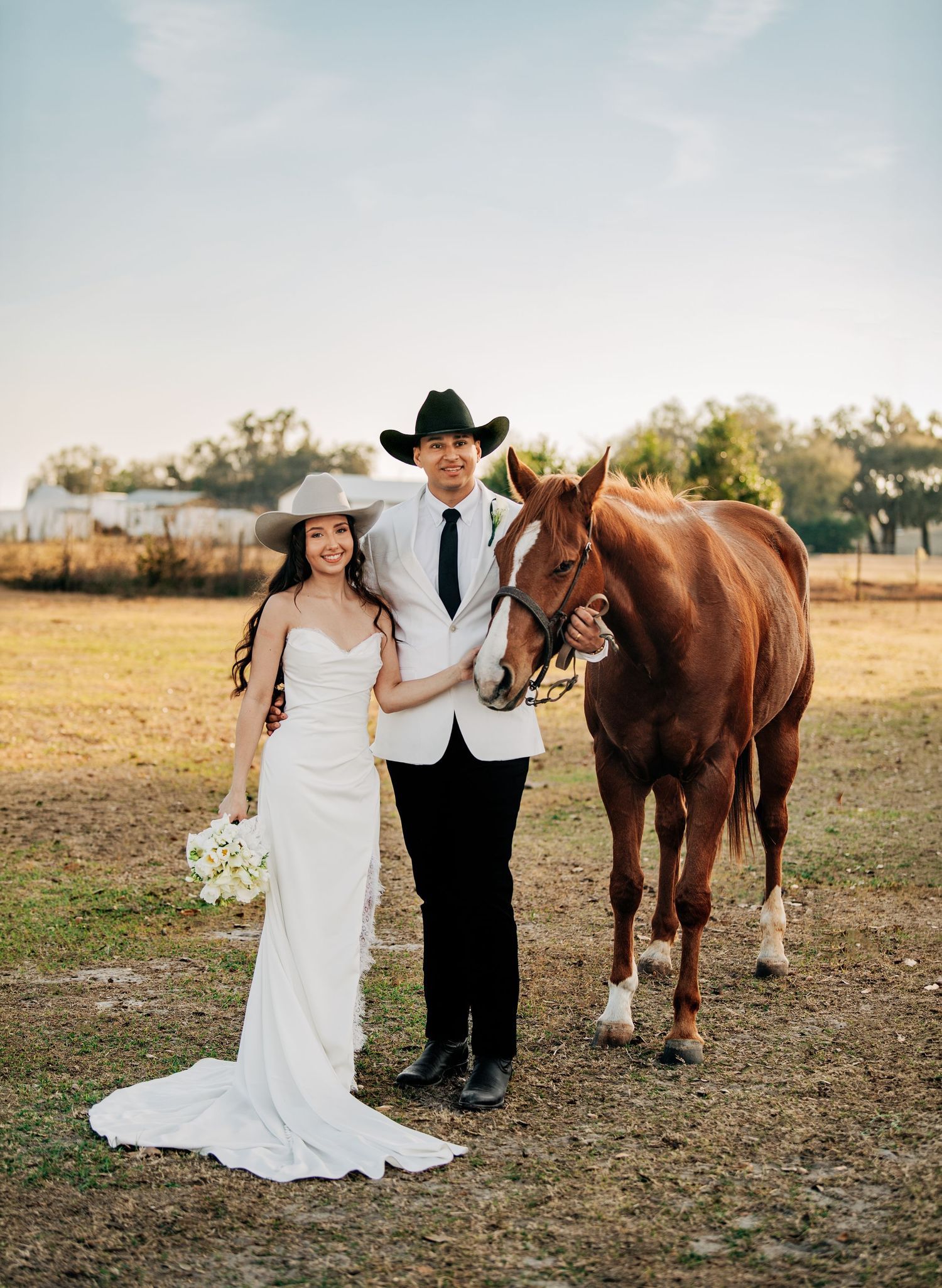 Bride and groom in cowboy hats pose with a chestnut horse on a ranch at golden hour.