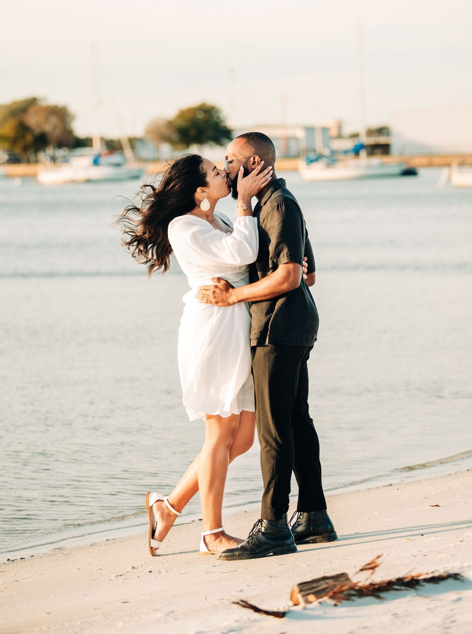 Couple kissing on a sunny beach, woman in white dress, man in dark outfit, sailboats in background.