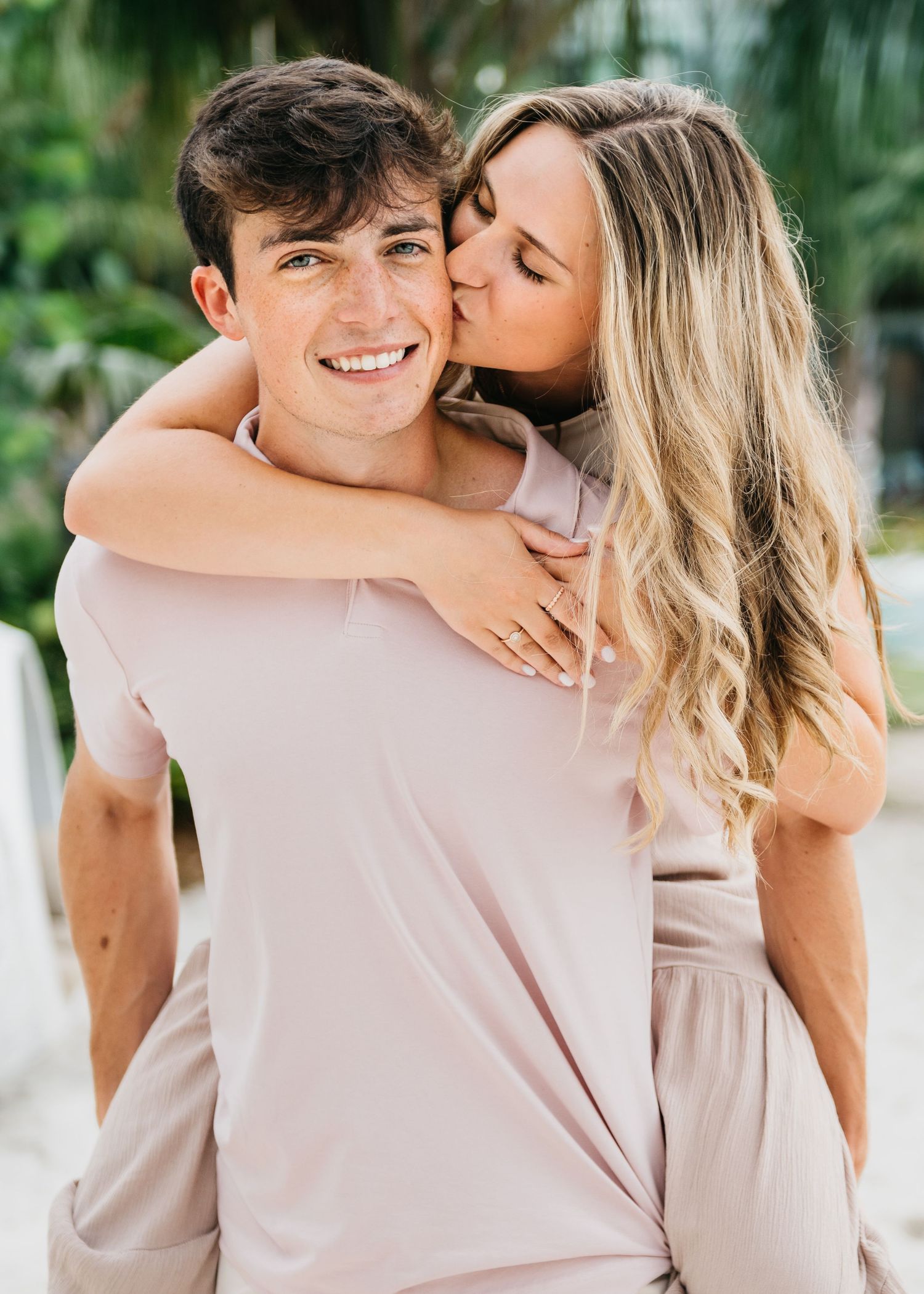 A smiling young man gives a piggyback ride to a blonde woman kissing his cheek, both wearing blush pink outfits.