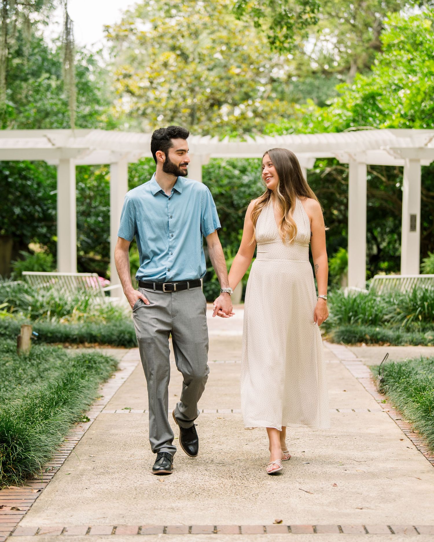 A couple walks hand-in-hand along a garden path, man in blue shirt and woman in white dress, surrounded by lush greenery.
