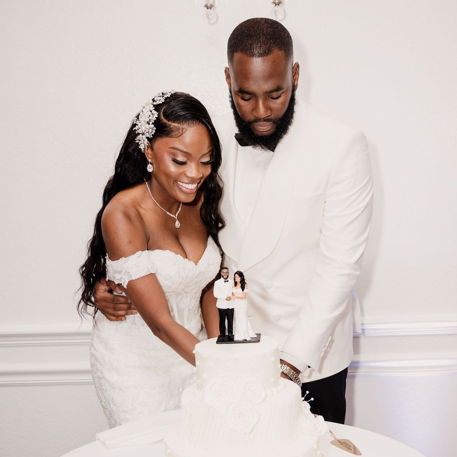 Bride and groom cutting wedding cake together, dressed in white wedding attire with floral cake topper.