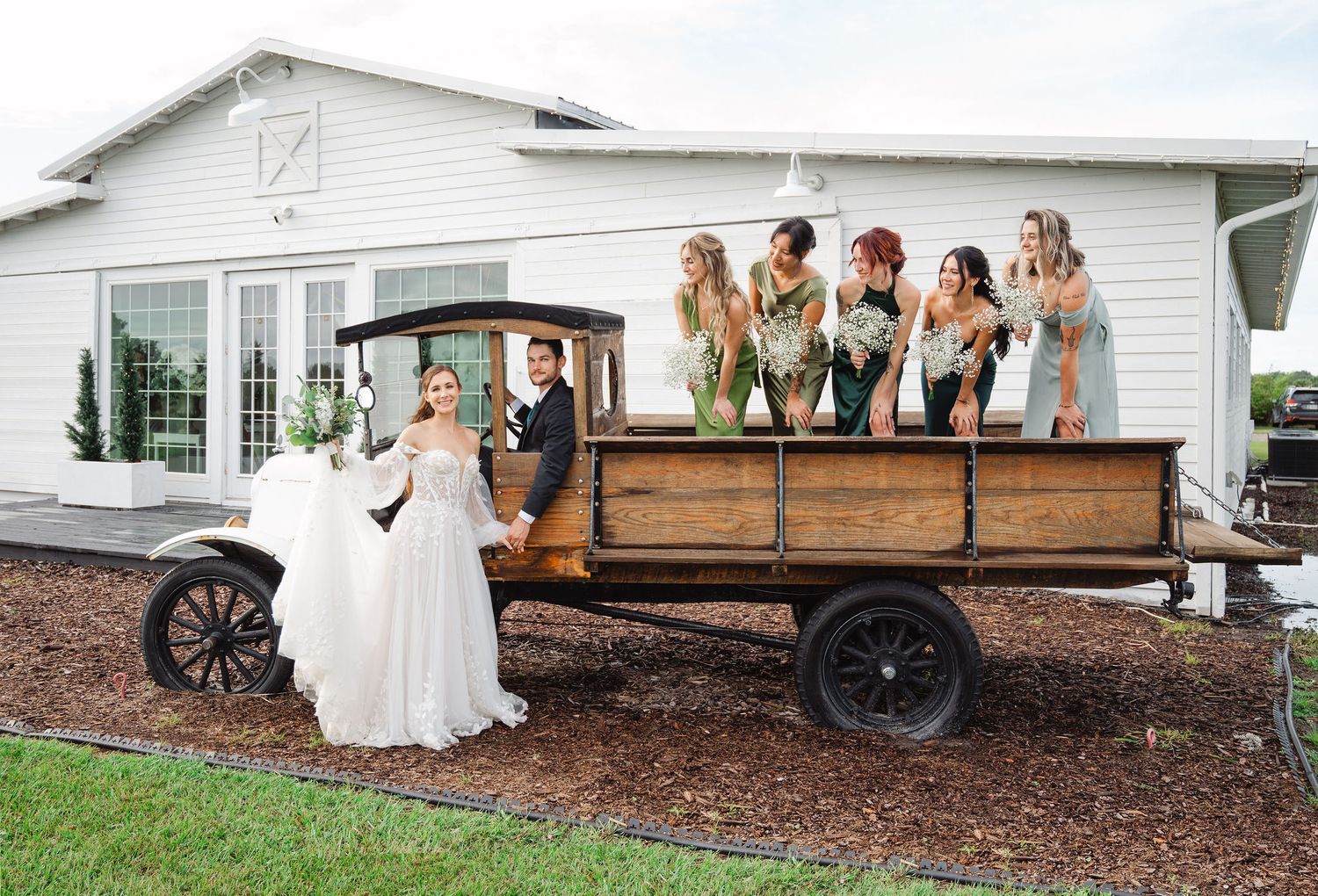 Bride and bridesmaids in colorful dresses posing with a vintage wooden flatbed truck outside a white barn venue.