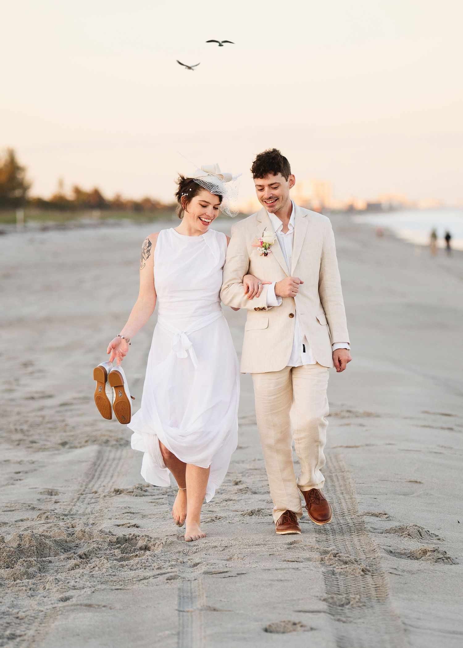 Bride in white dress and groom in tan suit walking barefoot on beach at sunset, smiling and holding shoes.