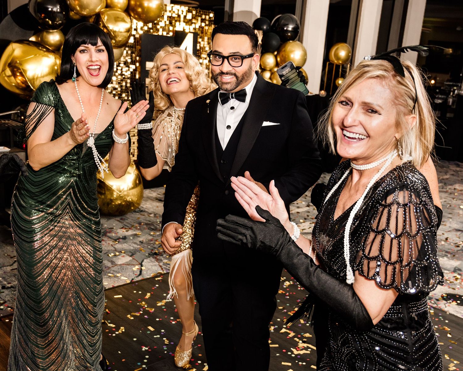 Four elegantly dressed guests celebrate at a glamorous black and gold New Year's Eve party with balloons and confetti.