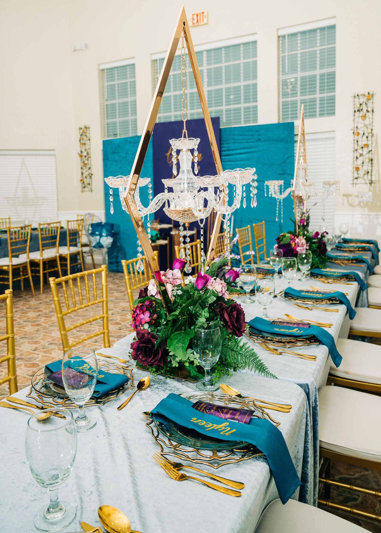 Elegant wedding reception table with gold chandelier centerpiece, teal napkins, gold flatware, and purple floral arrangements.