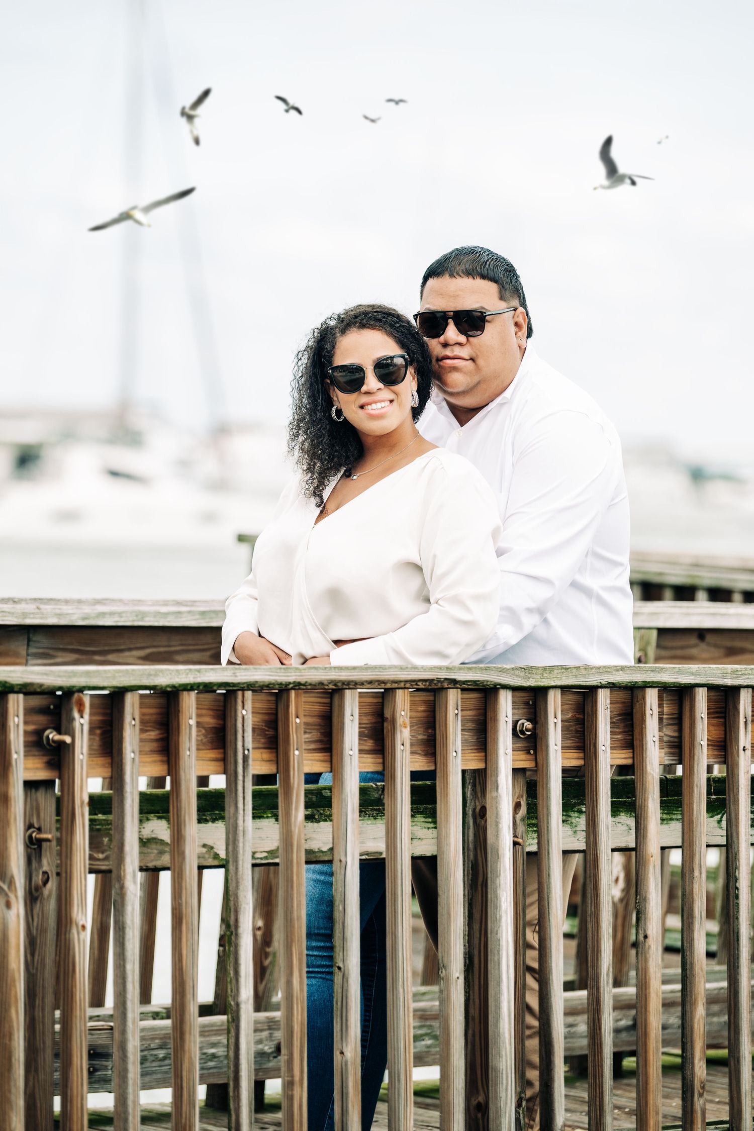 Couple in white tops and sunglasses smiling on a wooden pier with seagulls flying and sailboats in the background.