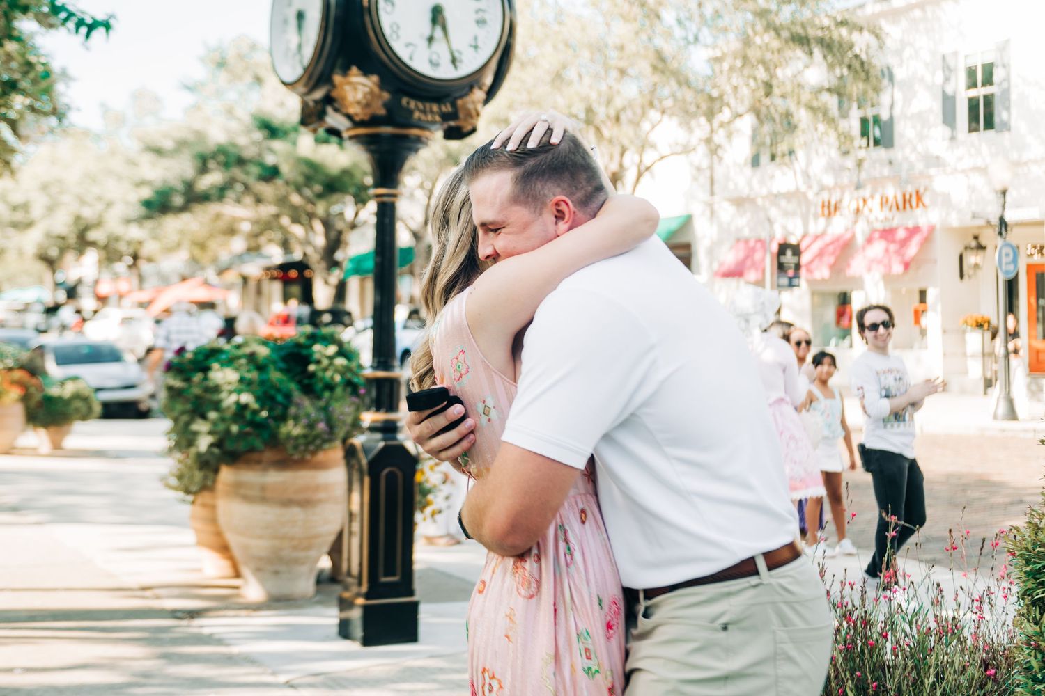 Couple embracing and hugging near a decorative street clock on a sunny outdoor sidewalk in a charming town.