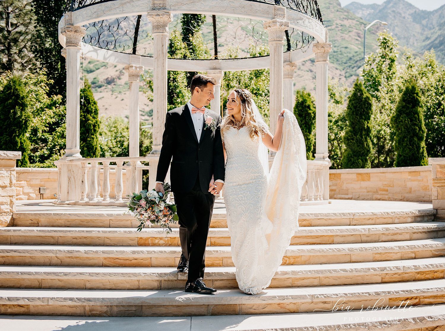 A bride and groom share a romantic moment on stone steps beneath a white gazebo with mountain views.