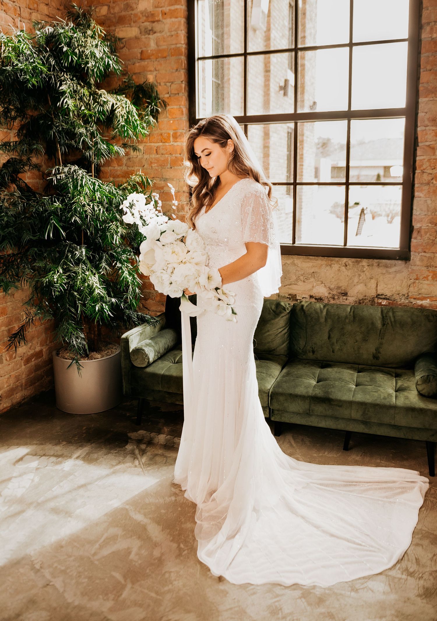 Bride in white lace gown with flutter sleeves holds white orchid bouquet near green velvet sofa and brick wall.