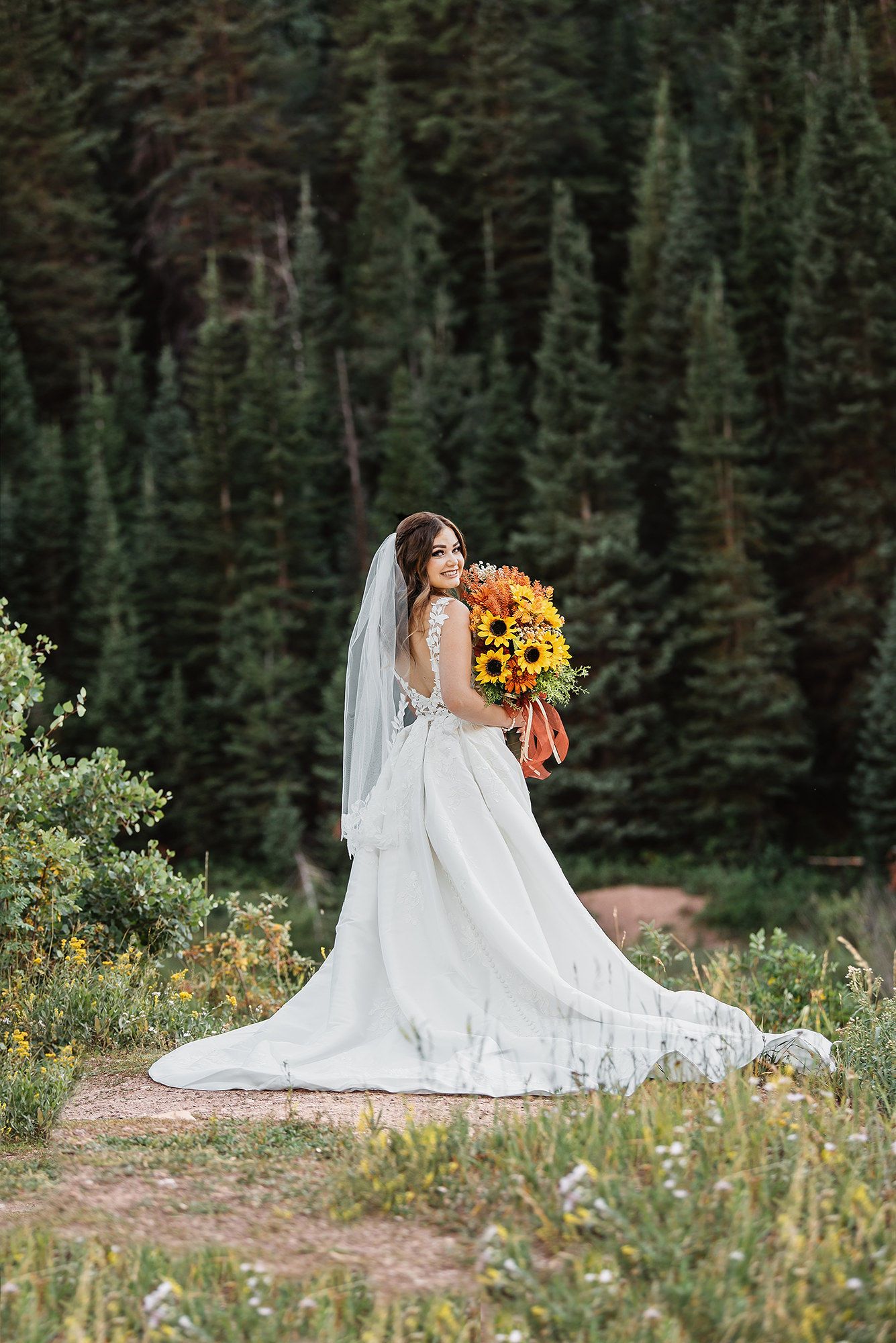 Bride in white gown with veil holds sunflower bouquet, standing before evergreen forest backdrop.