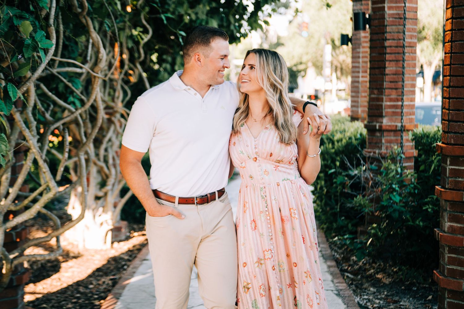 A couple smiles at each other while walking along a sunny brick pathway surrounded by lush greenery.