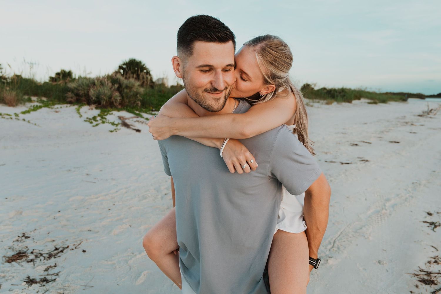 Couple enjoying a romantic piggyback ride on a sandy beach, smiling and embracing near lush greenery.