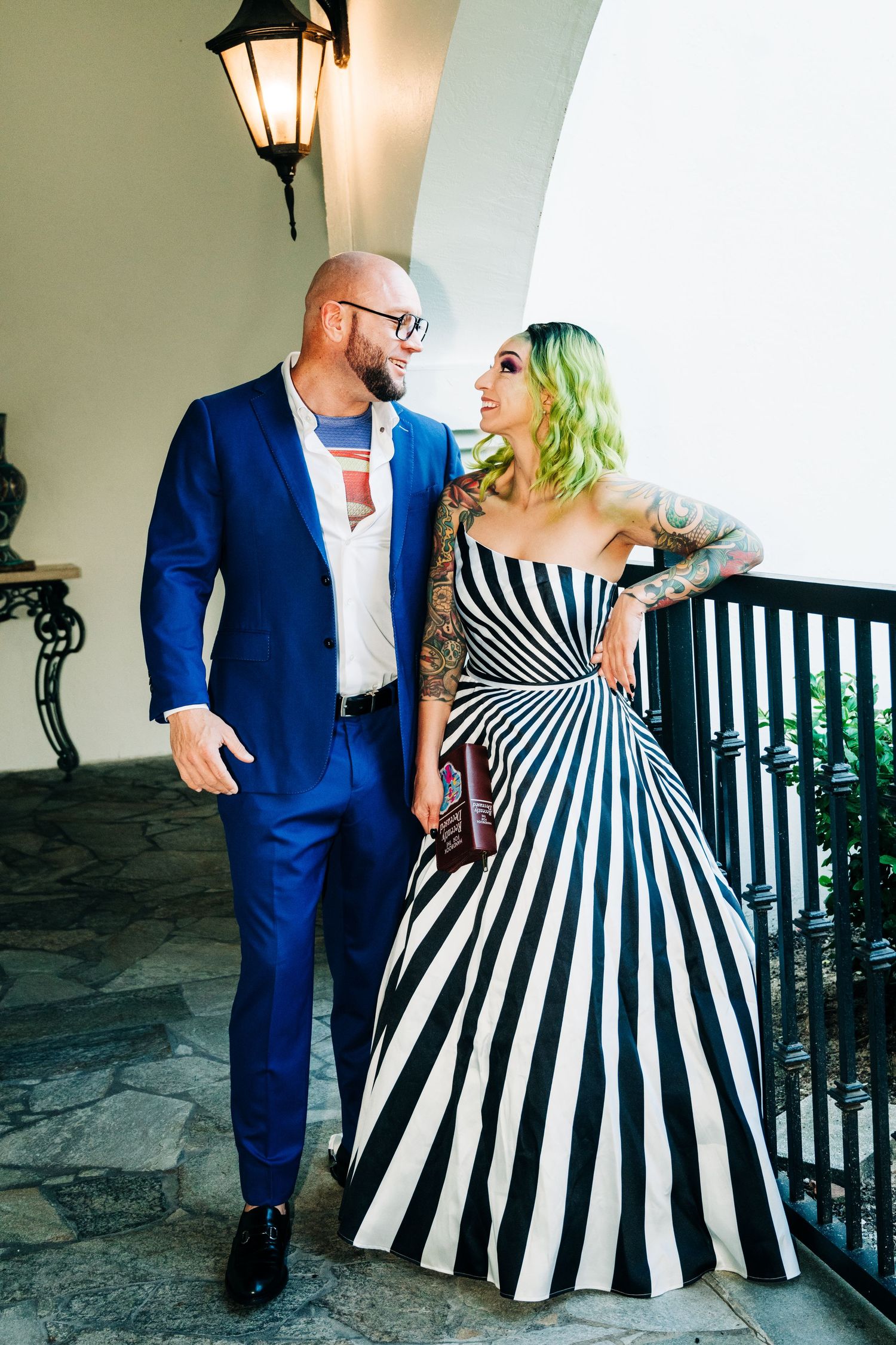 Couple posing on balcony, man in blue suit, woman in bold black and white striped strapless gown with tattoos.