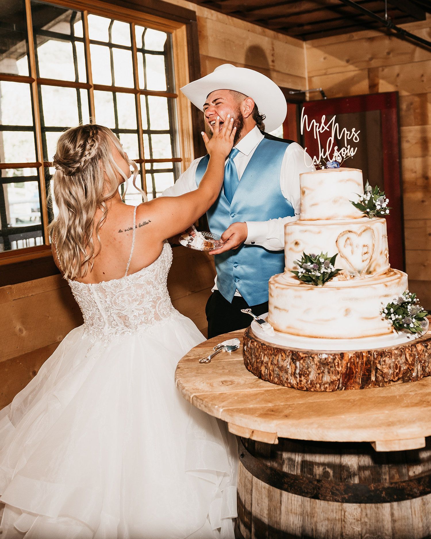 Bride smearing wedding cake on groom's face at rustic barn reception, with tiered cake on barrel stand.
