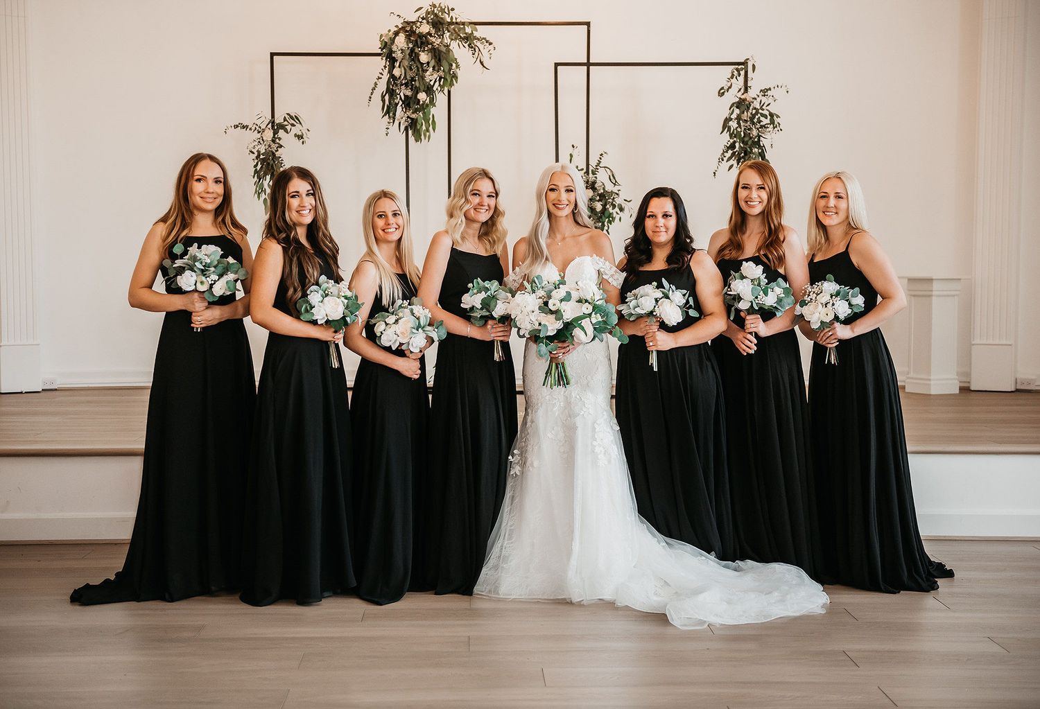 Bride in white gown with seven bridesmaids in black dresses holding white bouquets in elegant white venue.