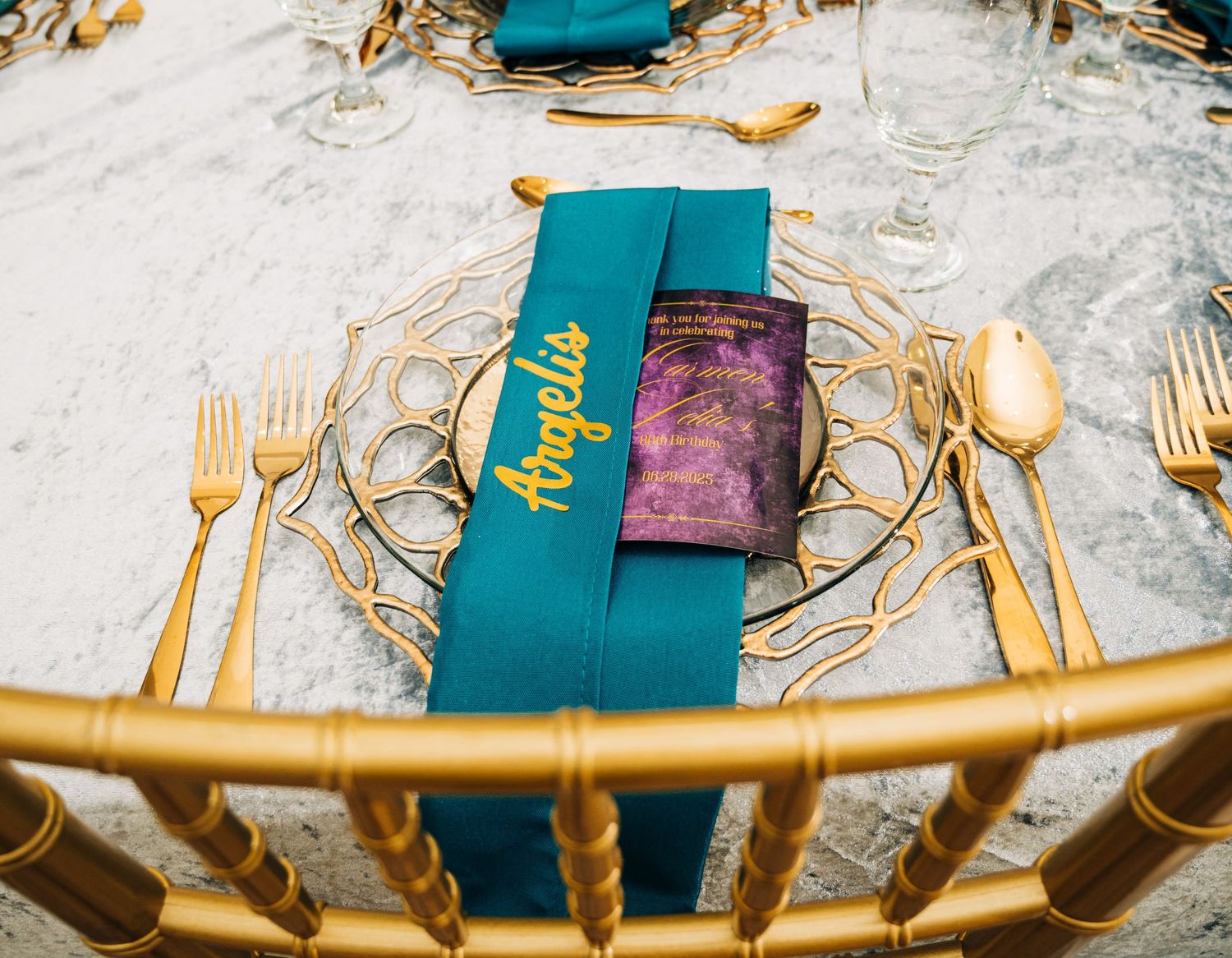 Elegant gold wedding place setting with teal napkin, purple menu card, and personalized name tag on marble table.
