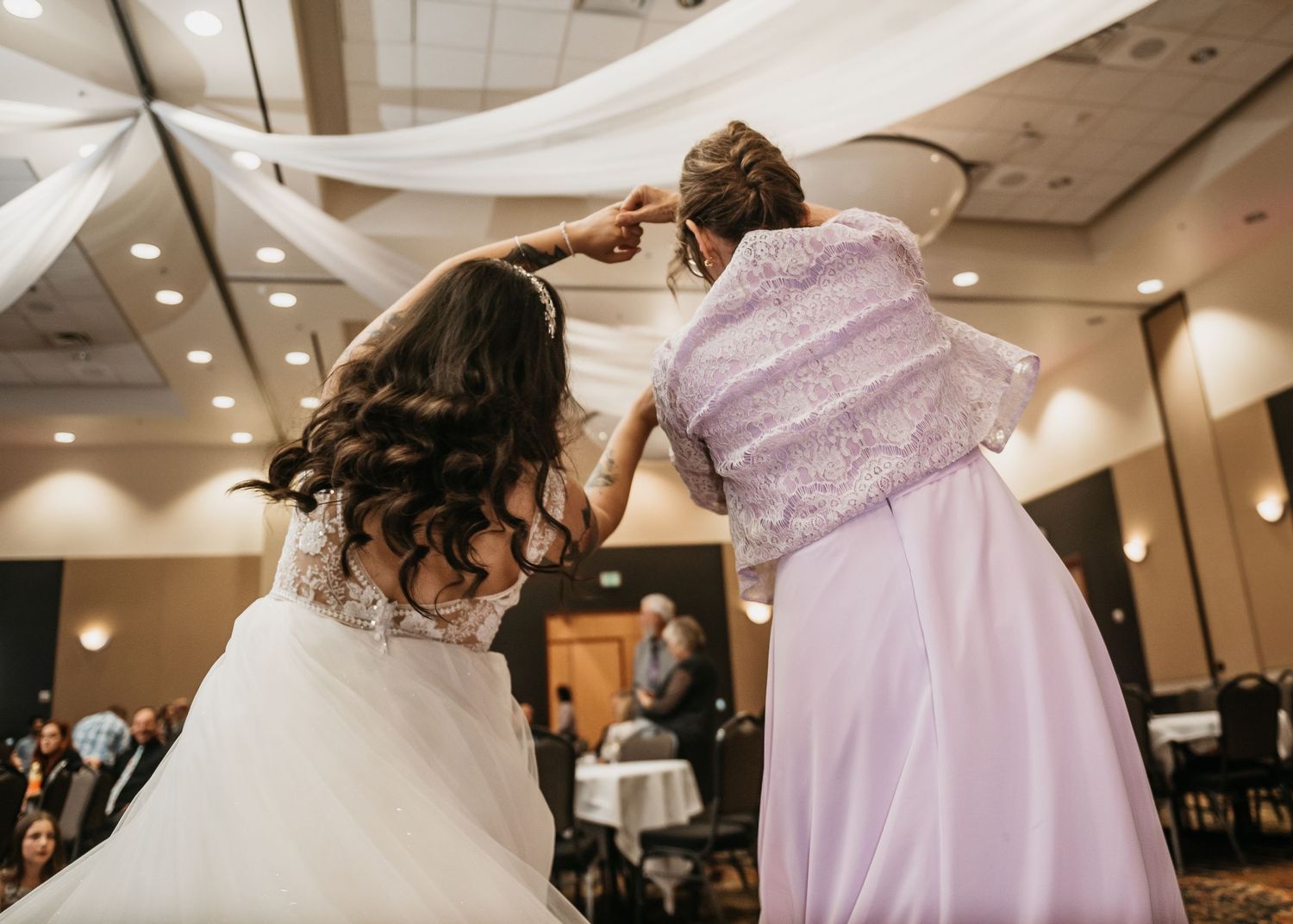 Bride and older woman in lavender gown share a dance at wedding reception in elegant ballroom.