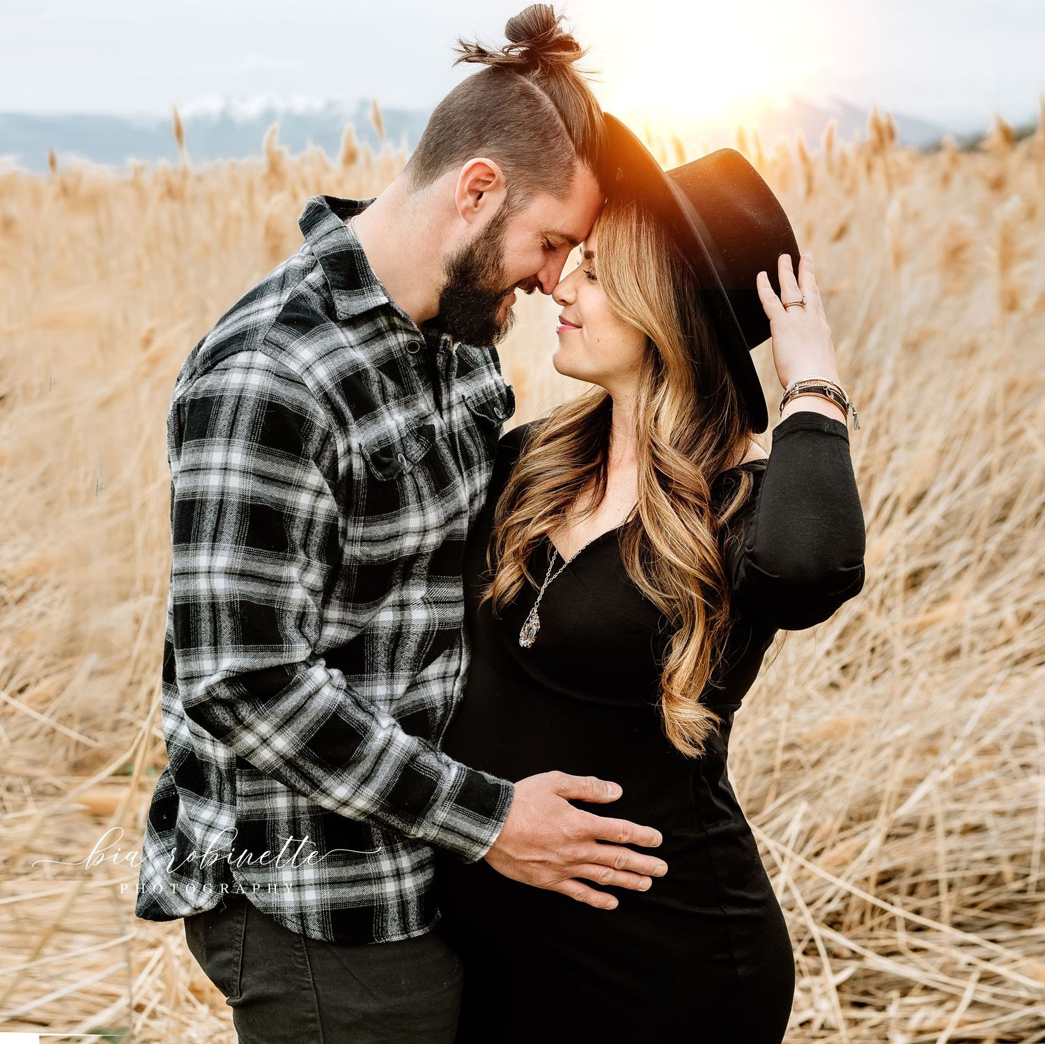 Couple sharing a romantic moment in a golden field at sunset, woman in black dress holding a hat, maternity photo session.