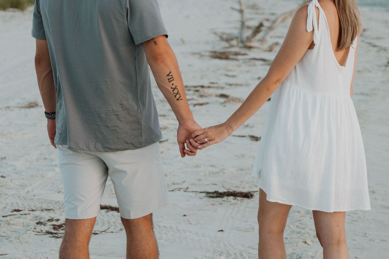 A couple holds hands on a sandy beach, the man with a tattoo wearing gray shirt and white shorts, woman in white dress.