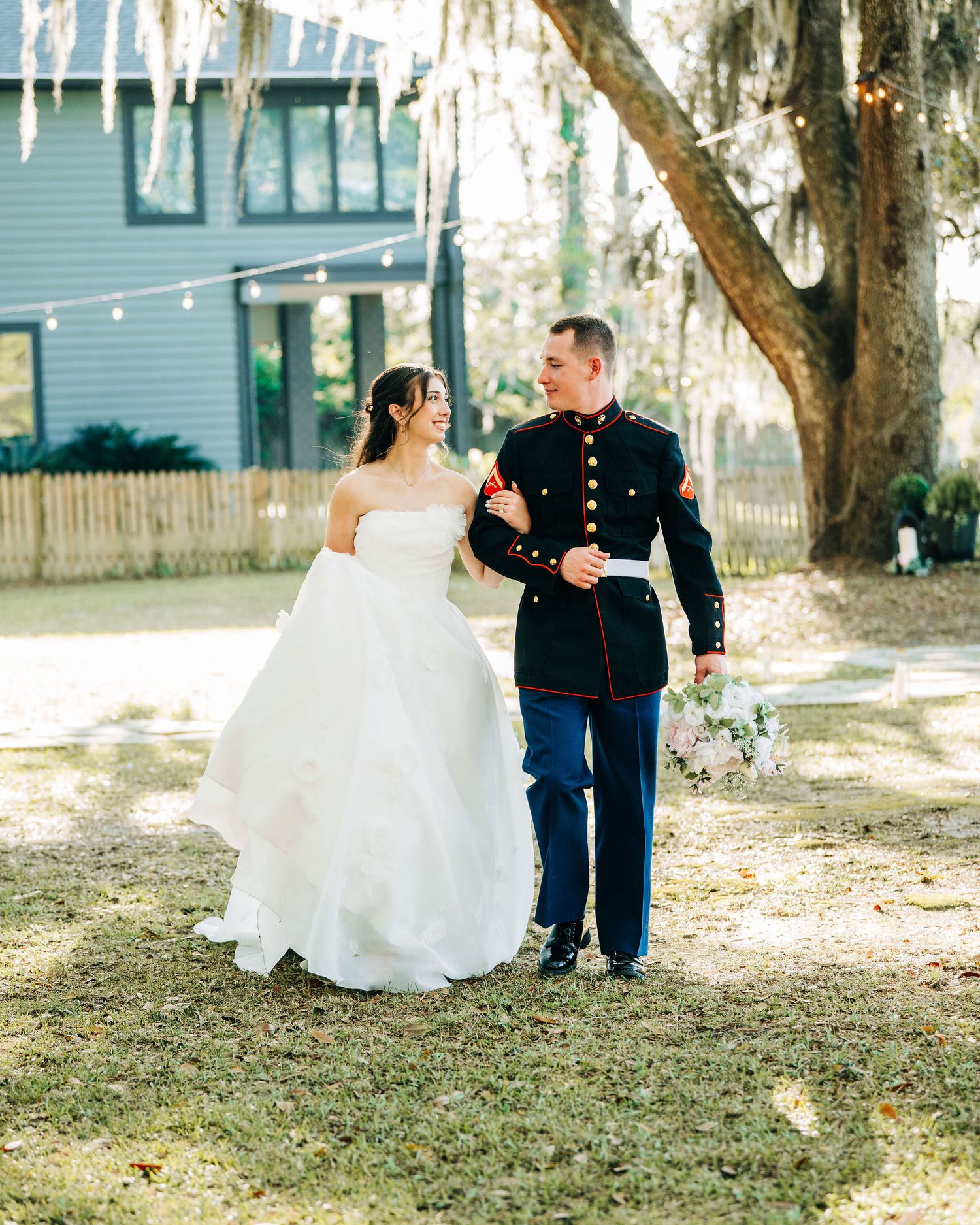 A bride in a white gown and groom in Marine dress blues walk together outdoors near a large oak tree.