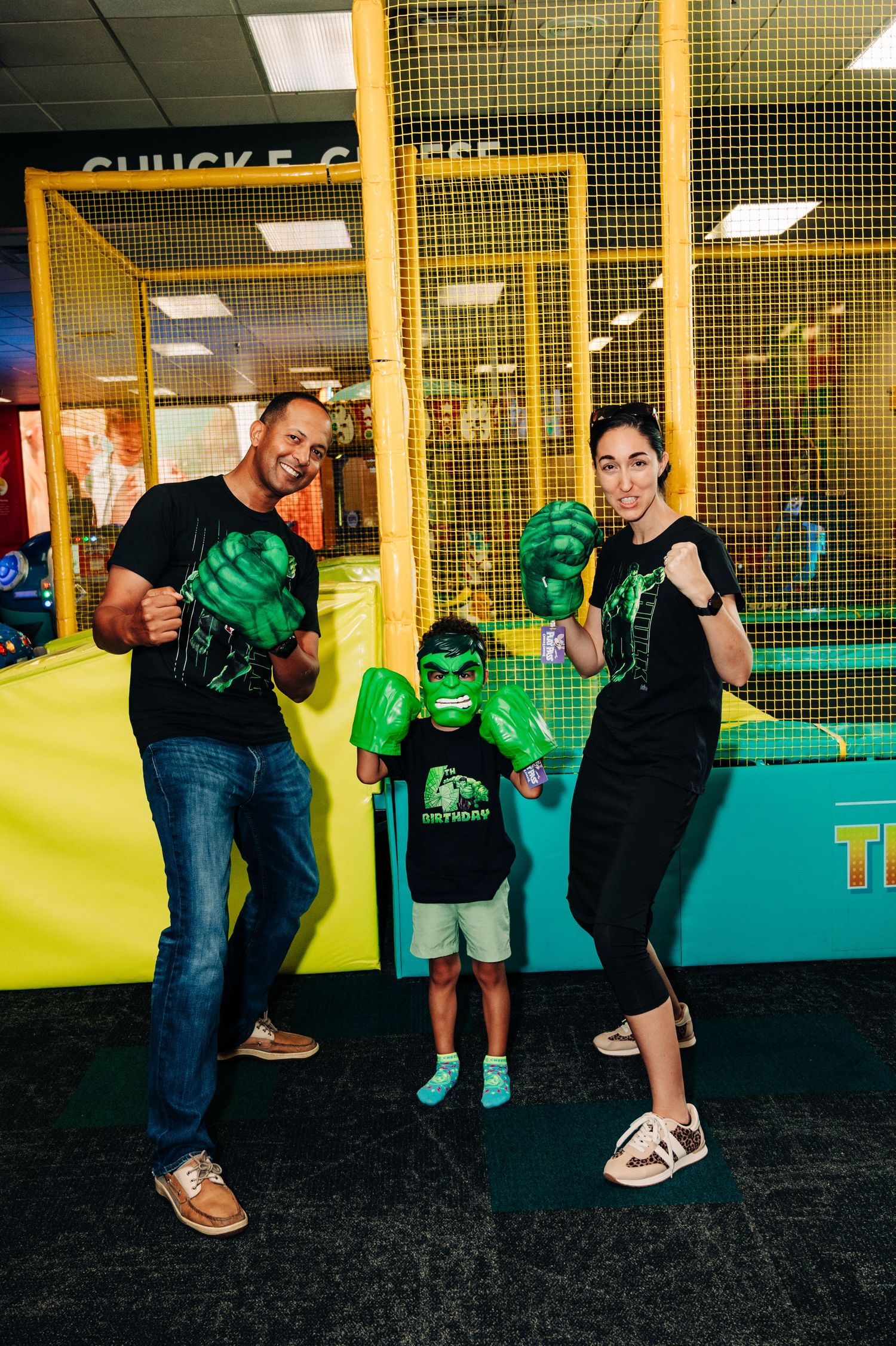 Family posing with Hulk-themed accessories at Chuck E. Cheese, wearing matching black Hulk t-shirts at a birthday party.