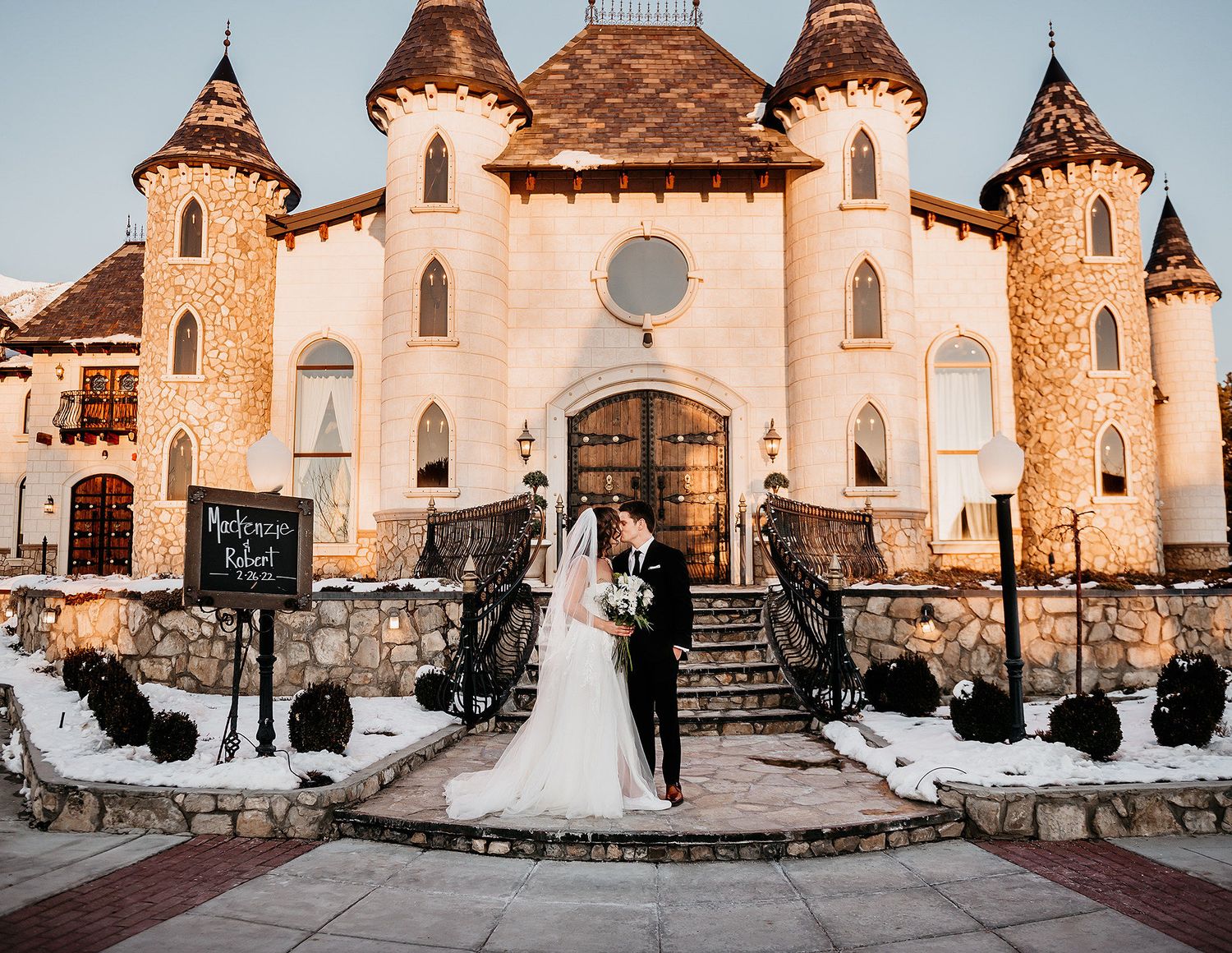 Bride and groom kiss in front of a fairy-tale castle wedding venue dusted with snow at golden hour.