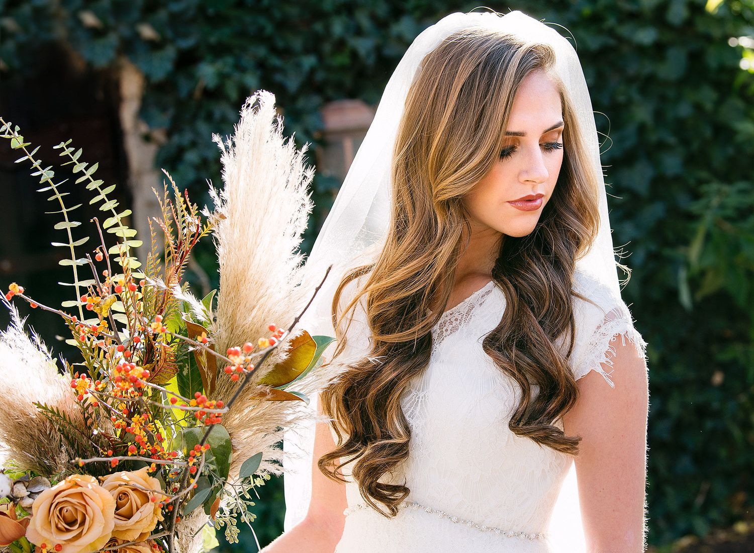 Bride with long wavy hair and veil holding a boho bouquet of pampas grass and roses in an outdoor setting.