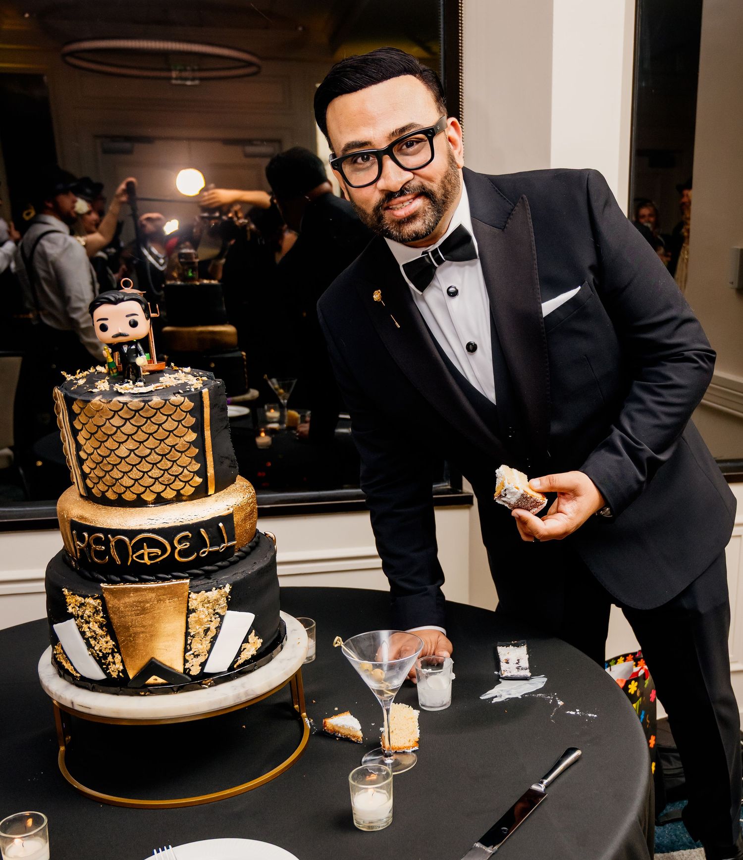 Man in black tuxedo smiling beside elegant black and gold tiered birthday cake with Mendell text at formal event.