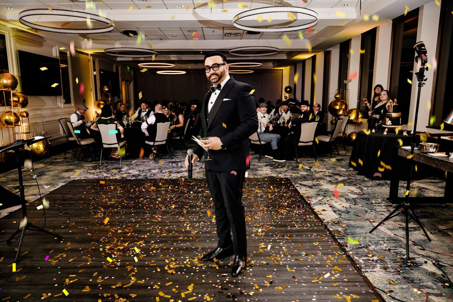 Man in black tuxedo stands on confetti-covered floor at elegant event venue with guests seated in background.