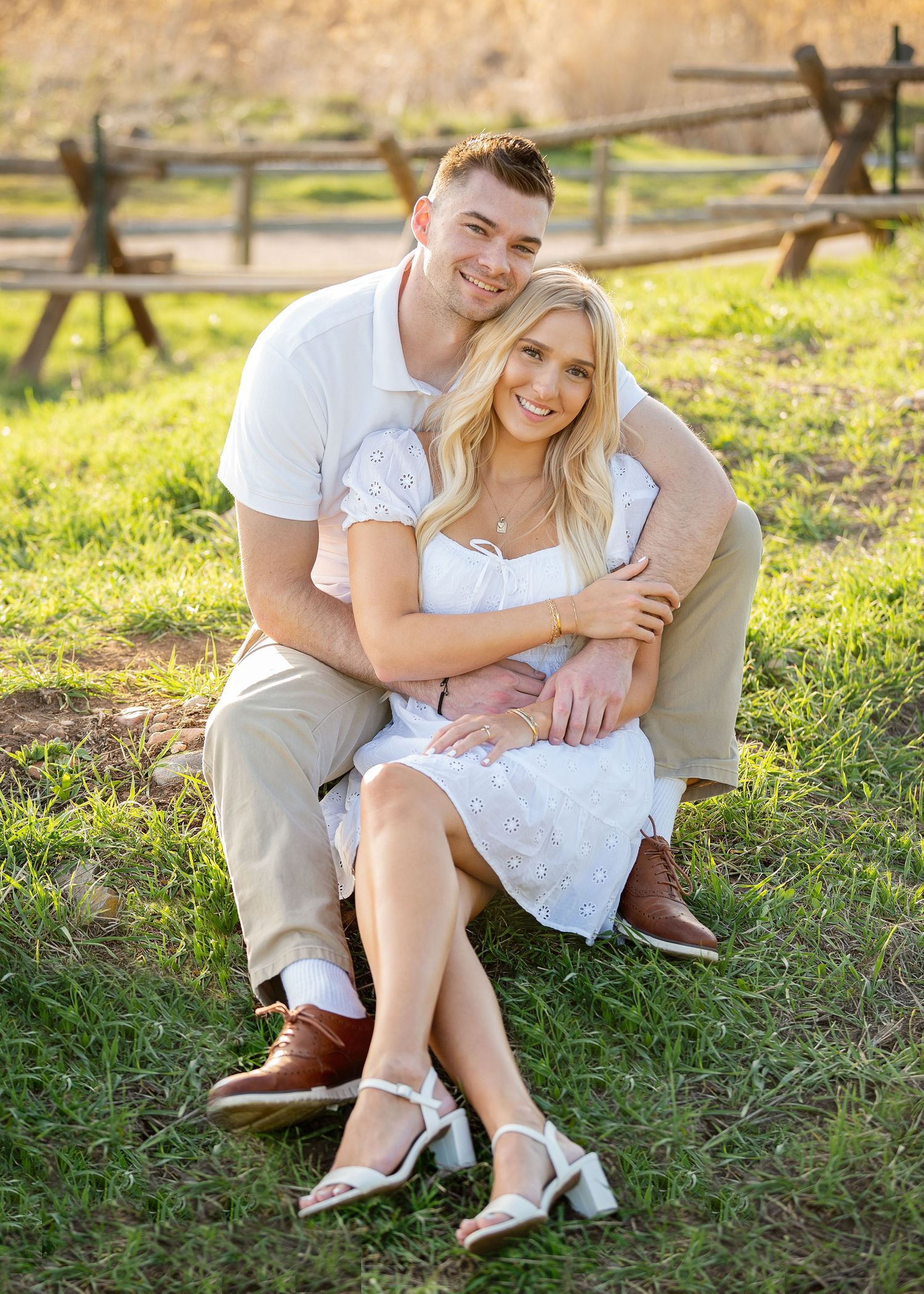 Happy couple sitting on grass in golden hour light, woman in white dress, man in khaki, with rustic fence backdrop.