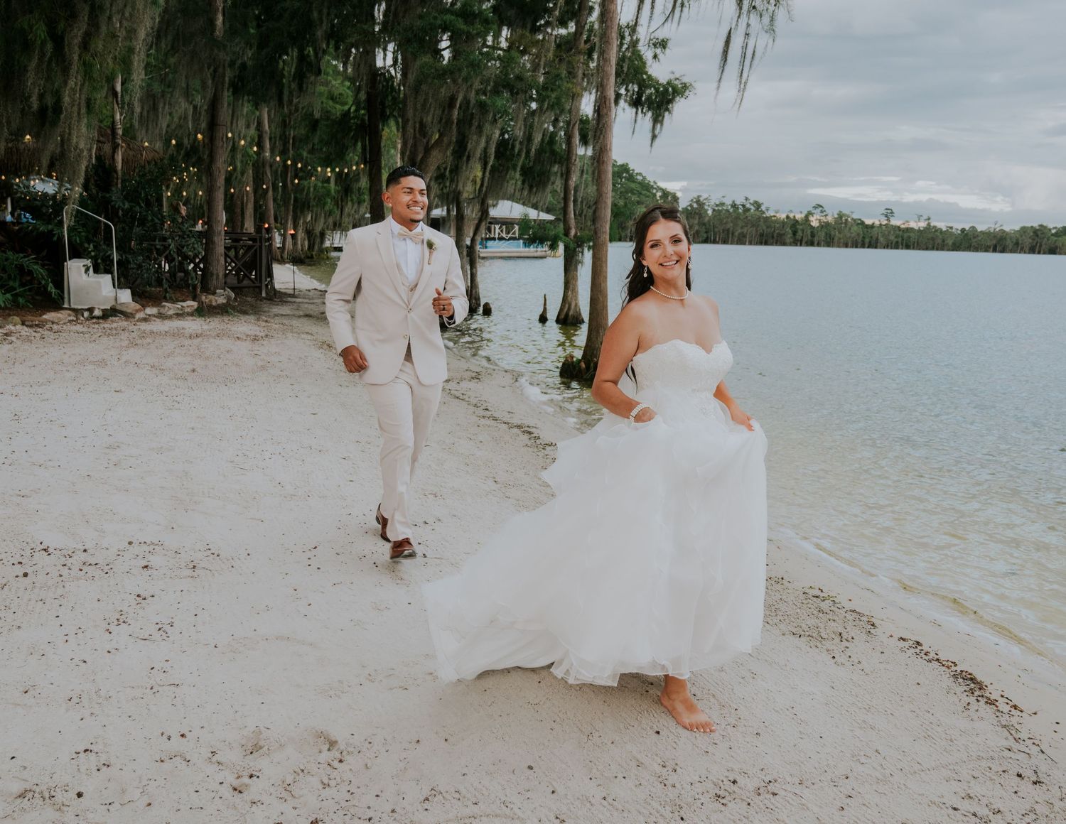 A joyful bride and groom walk hand-in-hand along a sandy lakeside beach surrounded by moss-draped trees.