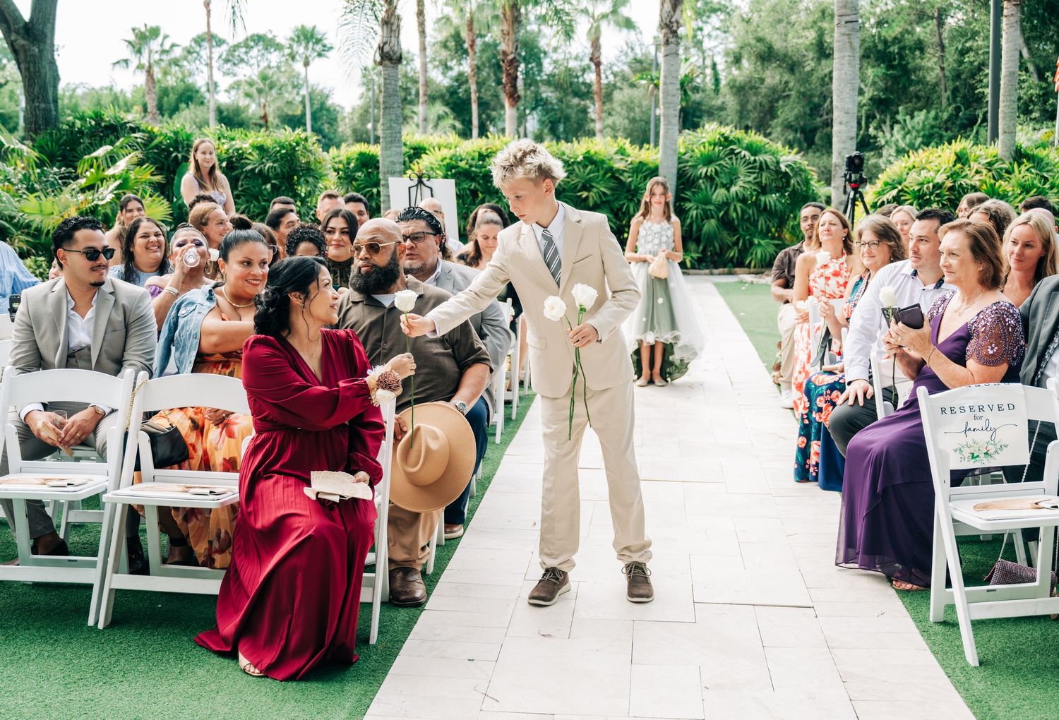 A groom walks down the aisle at an outdoor wedding ceremony, greeting guests seated in white chairs.