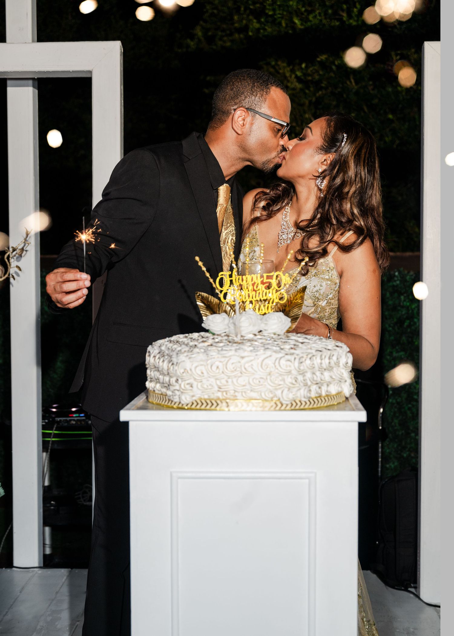 Couple kissing over elegant white and gold wedding cake with sparklers at romantic outdoor celebration.
