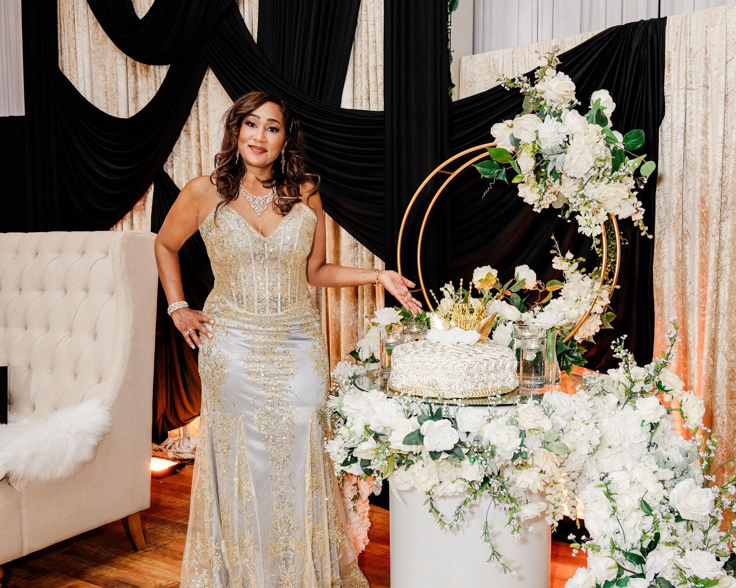 Woman in gold gown poses beside elegant white cake with floral arrangements at a glamorous celebration event.