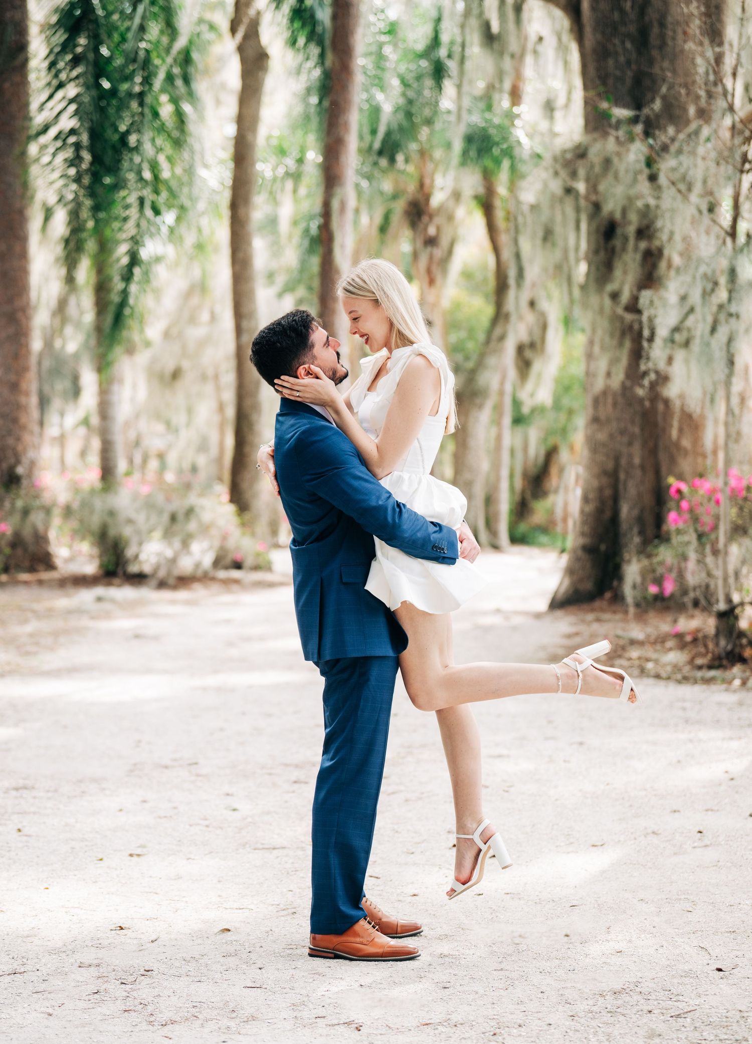 A man in a blue suit lifts a woman in a white dress and heels for a romantic kiss on a tree-lined path.