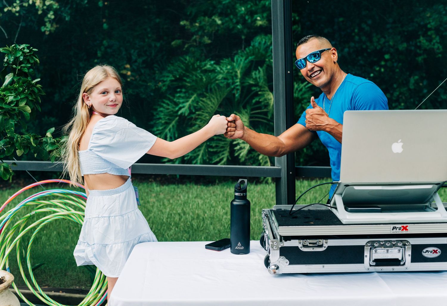 A young girl fist bumps a smiling DJ at an outdoor event setup with a laptop and equipment on a white table.